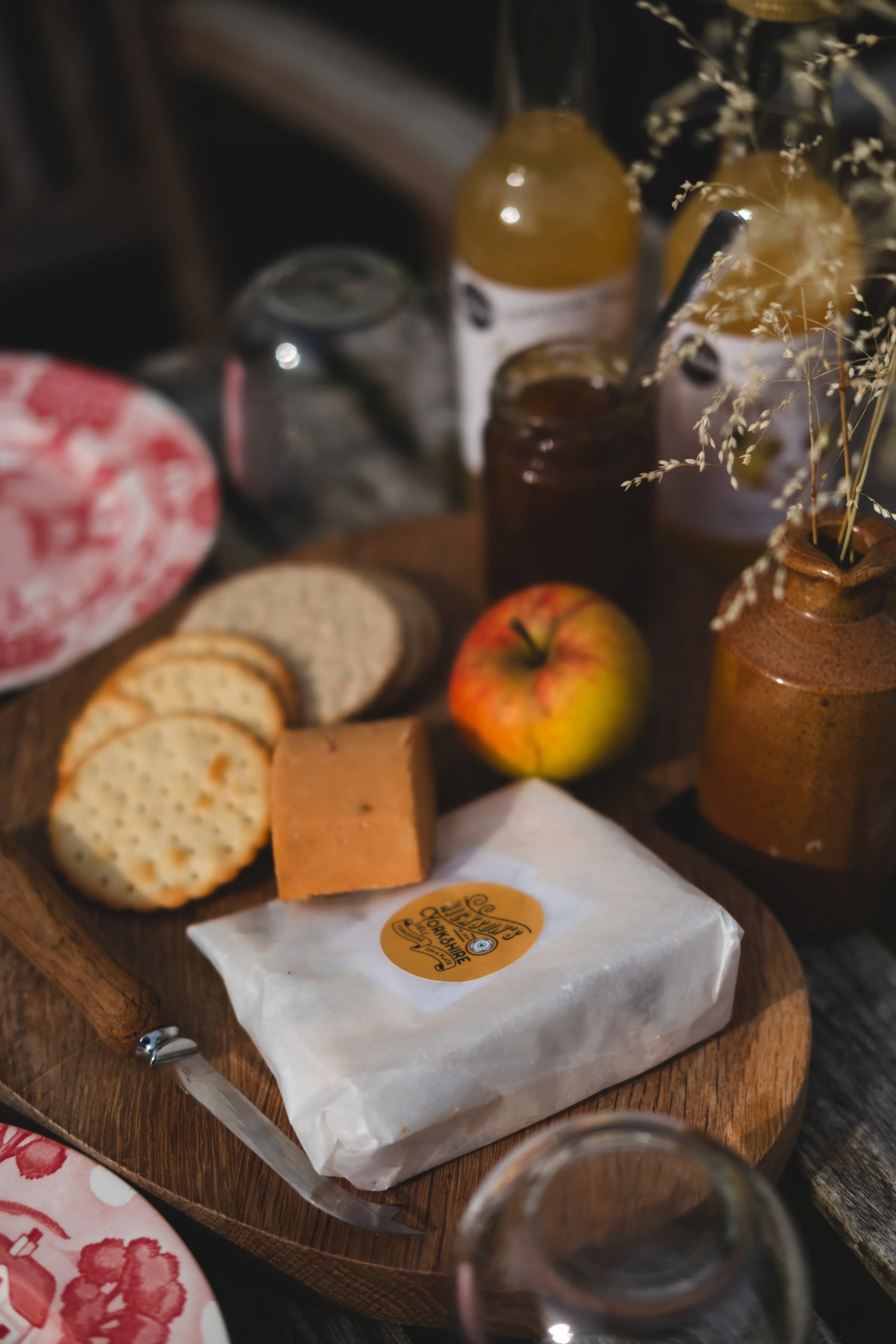 A cheese platter with crackers, a block of cheese, an apple, and jars of jam or honey, on a wooden cutting board.