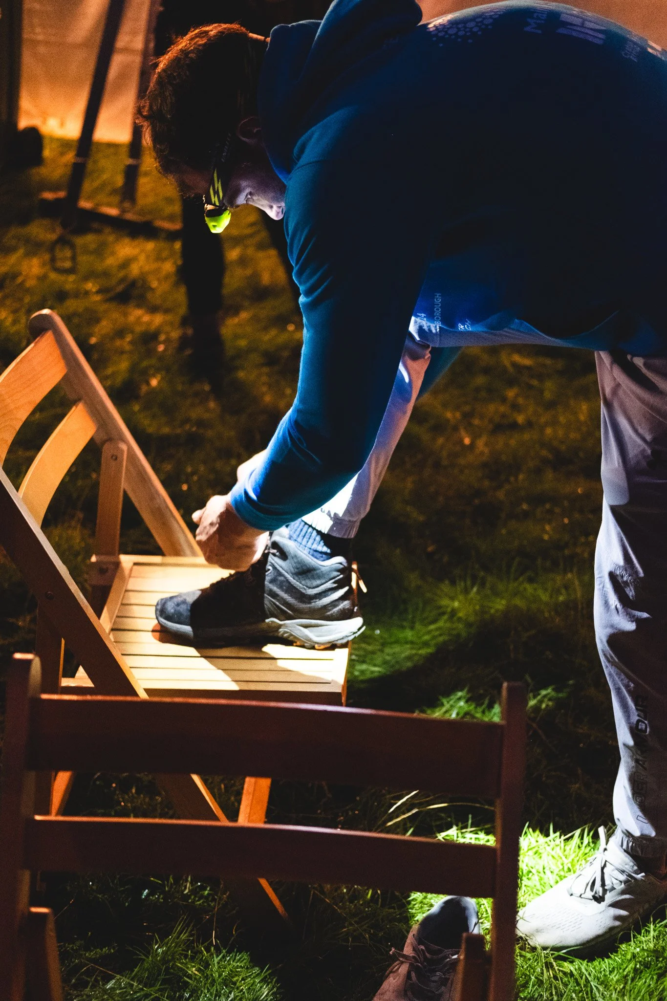 A person putting on their shoes, illuminated by a headlamp at night outdoors.