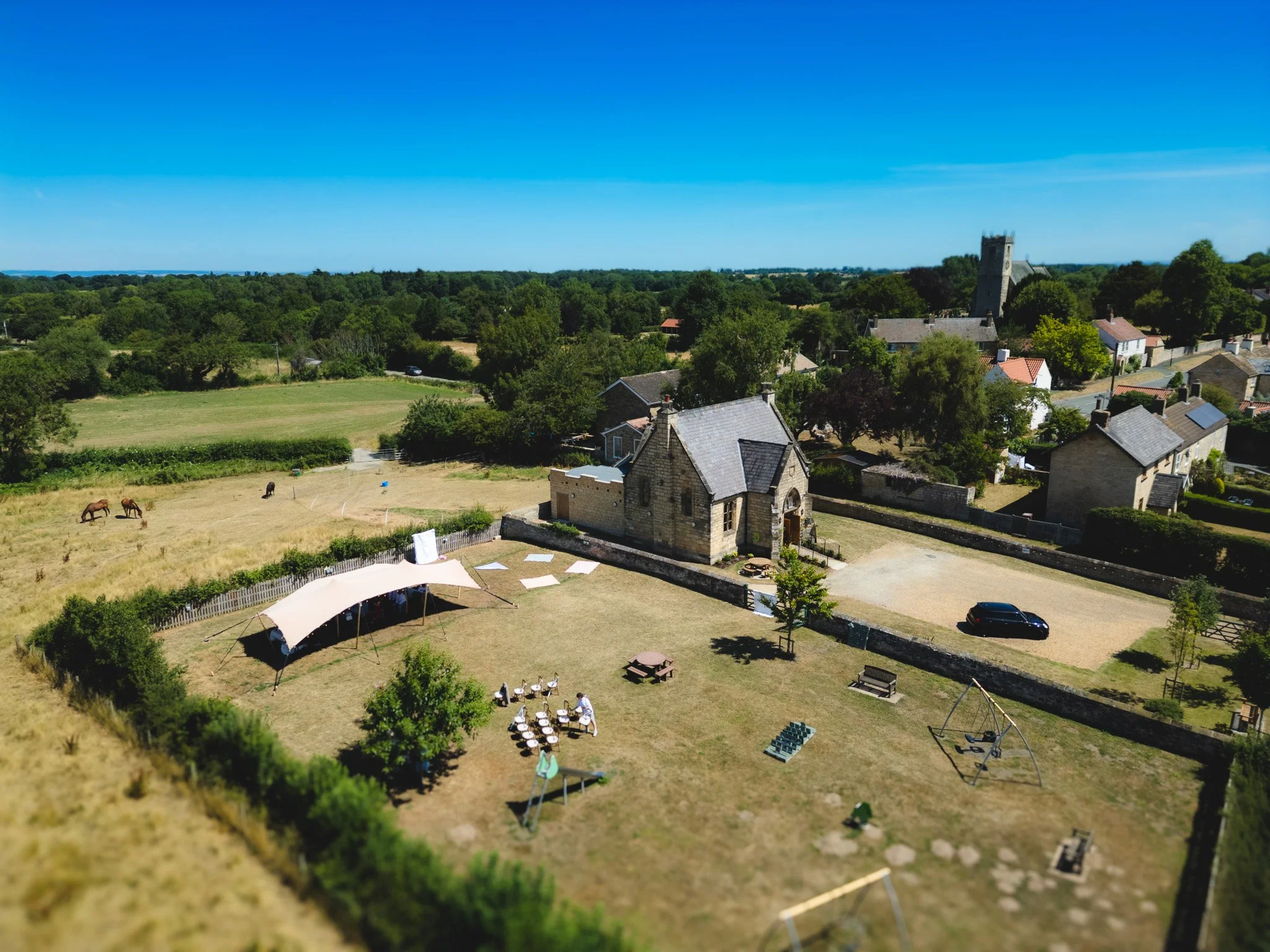 Aerial view of a church and grassy outdoor area with tents, lounge chairs, a playground, trees, and a woman with children. Nearby houses and a church tower in the background.