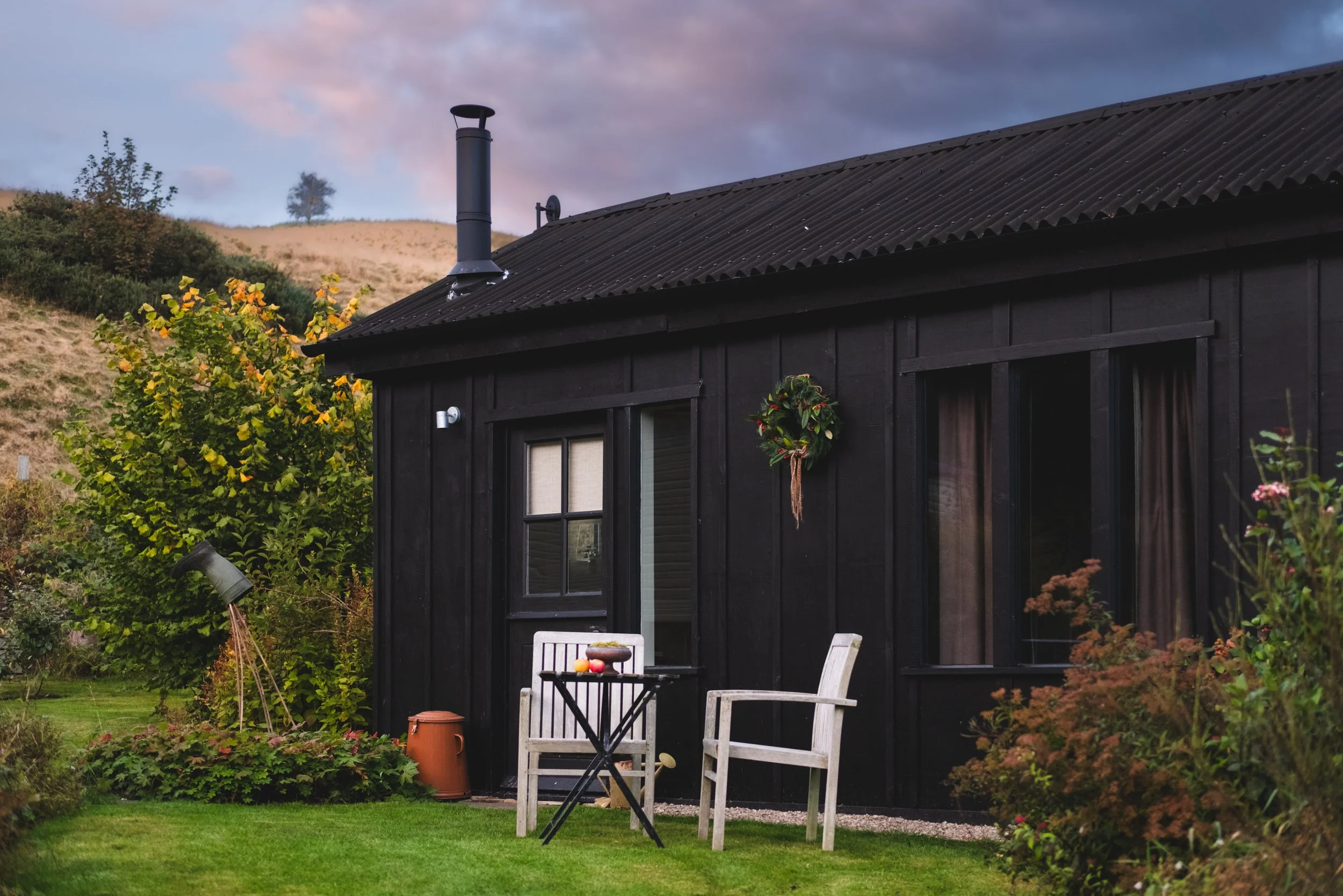 Black wooden house with a wreath on the wall, patio furniture, and garden plants in a green yard at dusk.