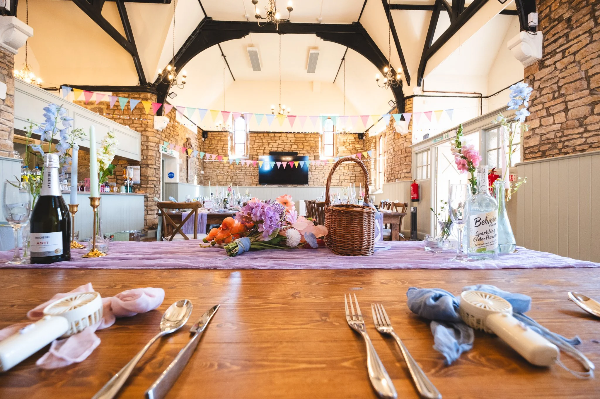A decorated banquet hall with brick walls, colorful bunting, flowers, candles, and set tables for a celebration or event.