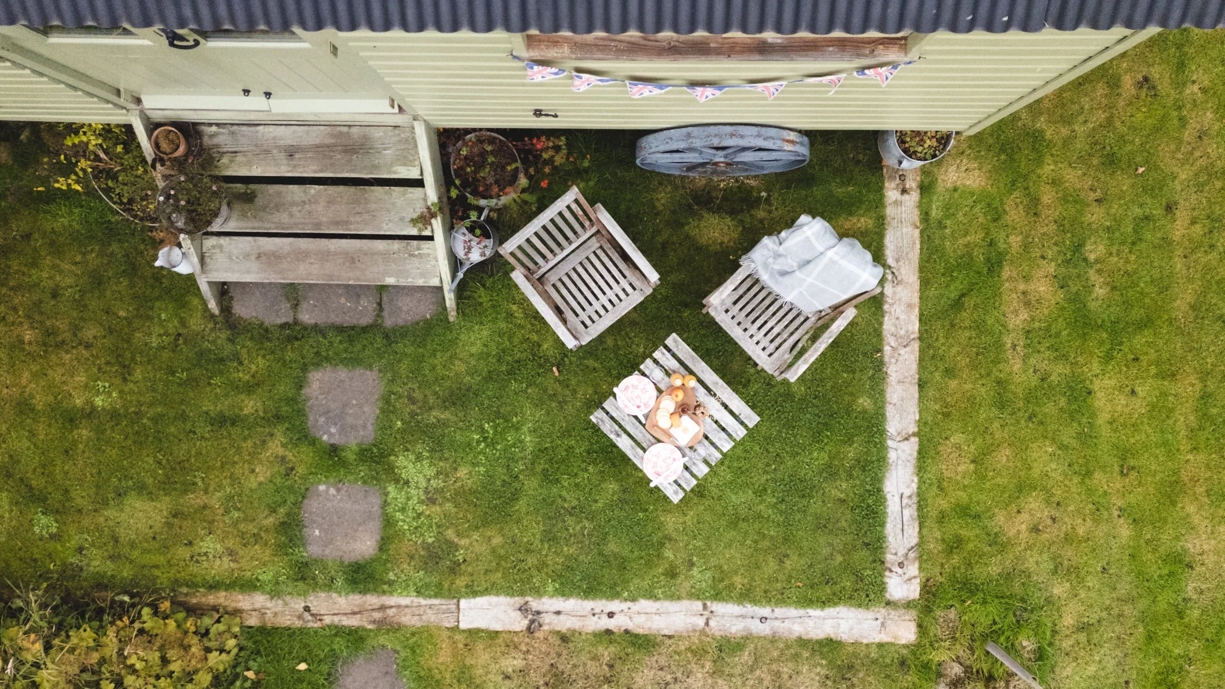 A top-down view of a small backyard patio with wooden chairs, a small table set with cups and a fruit bowl, surrounded by potted plants and a grassy lawn. The patio is bordered by a stone path and a wooden fence with bunting hanging on the fence.