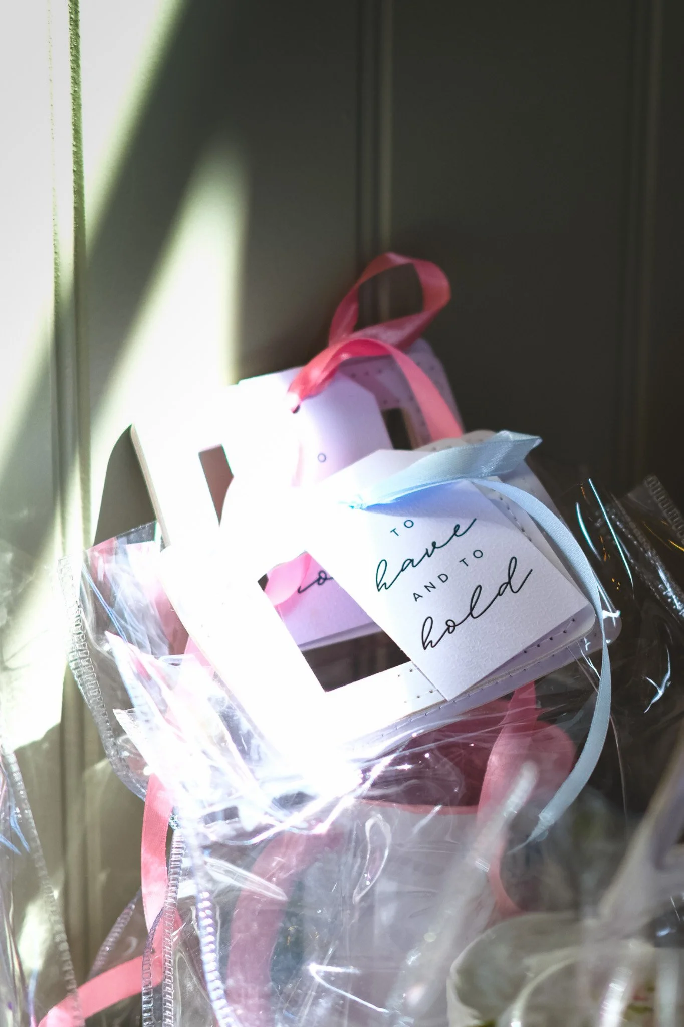 A small gift box wrapped with a pink ribbon, labeled 'To have and to hold,' resting on a shelf with sunlight casting shadows.