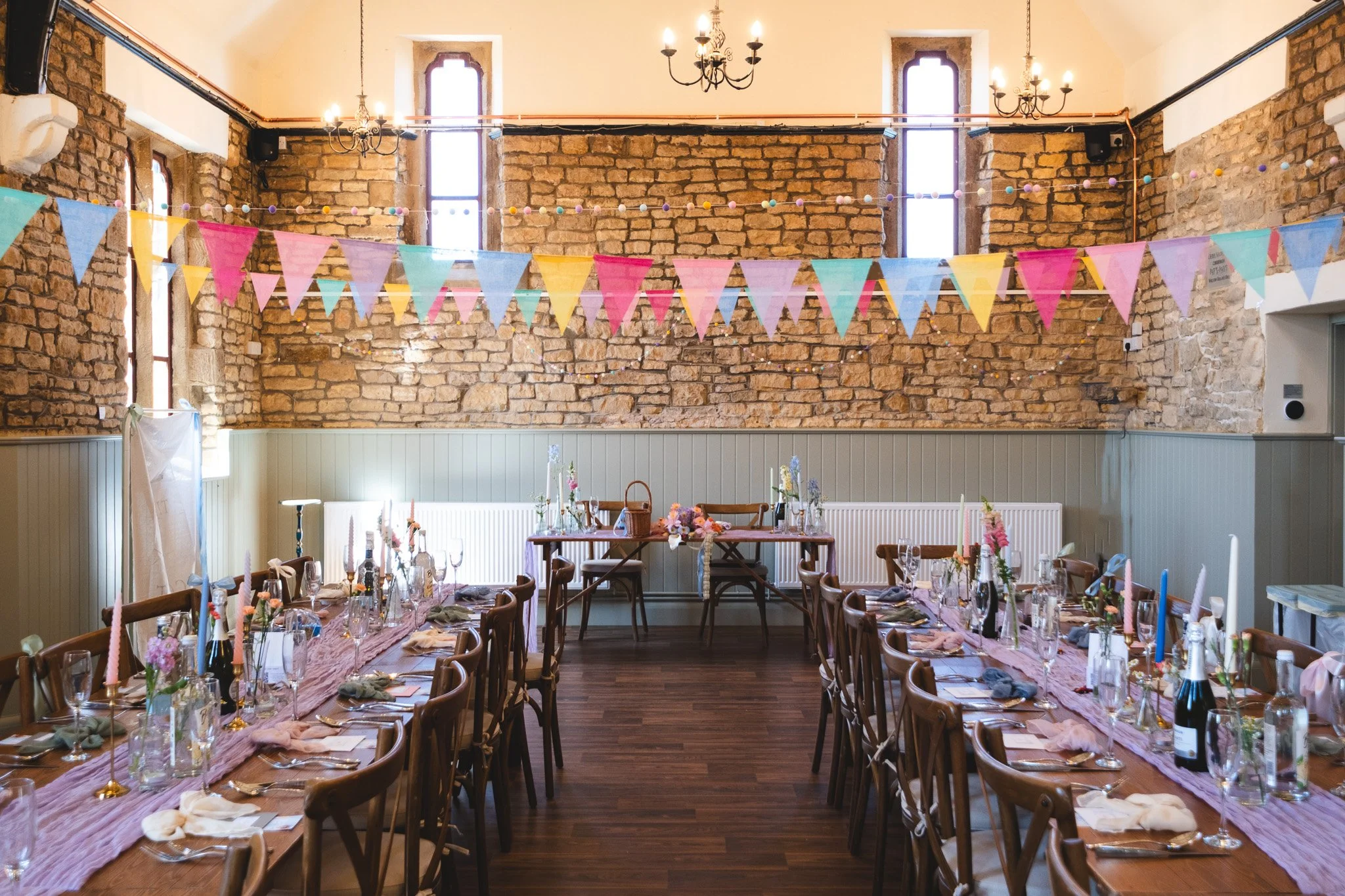 A decorated banquet hall with two long tables set for a celebration, adorned with pink, purple, and gold accents, floral centerpieces, and pastel bunting across the brick wall in the background.