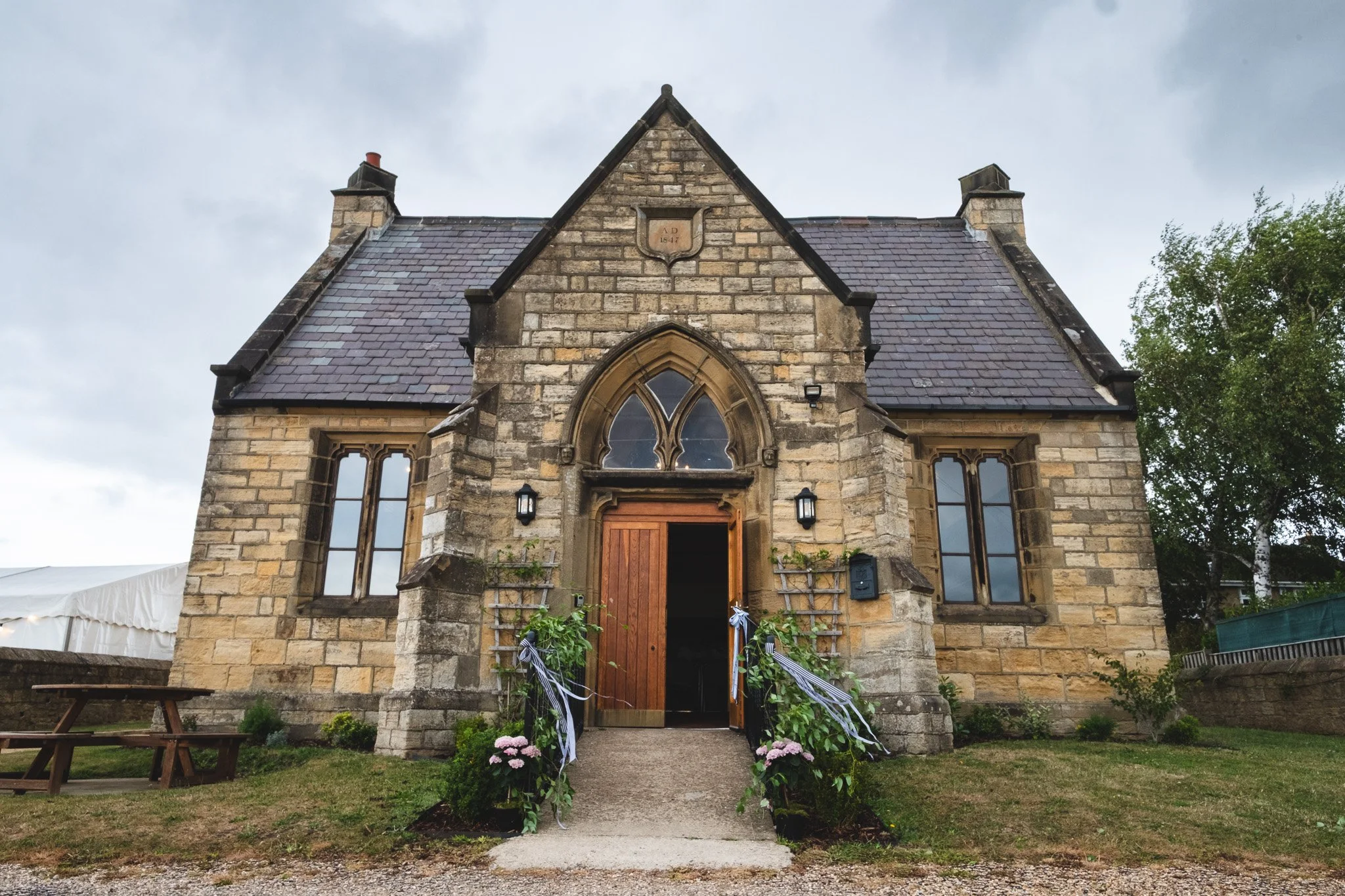 A historic stone church decorated for a wedding, featuring an open wooden door, arched windows, and floral arrangements with ribbons at the entrance.