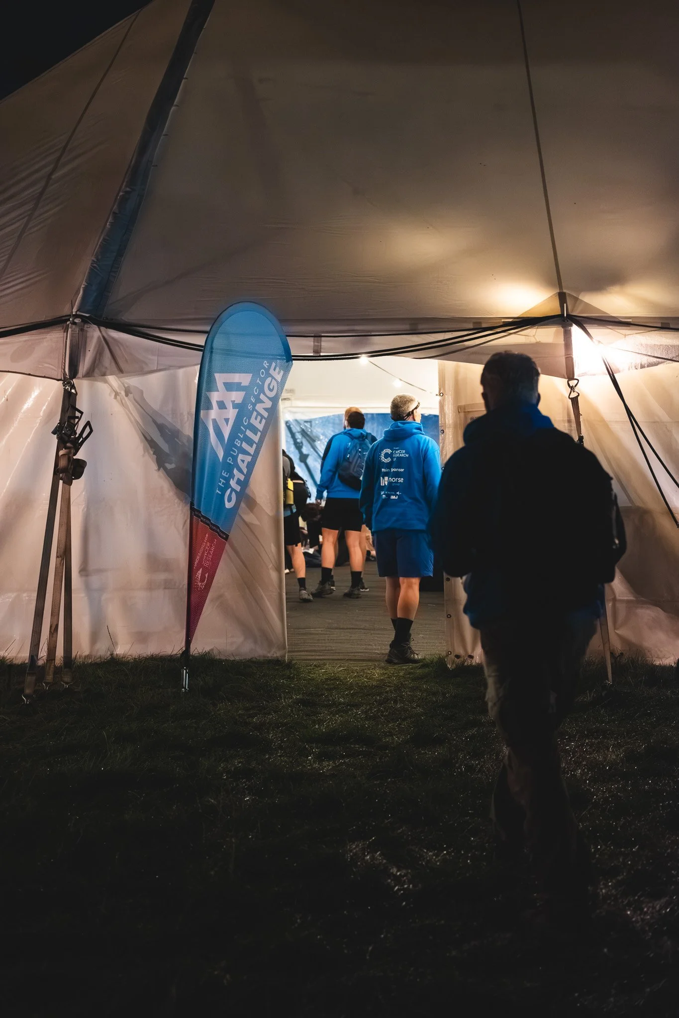 People entering a tent at night during a challenging outdoor event, with a blue banner that reads 'The Publicator Challenge'.