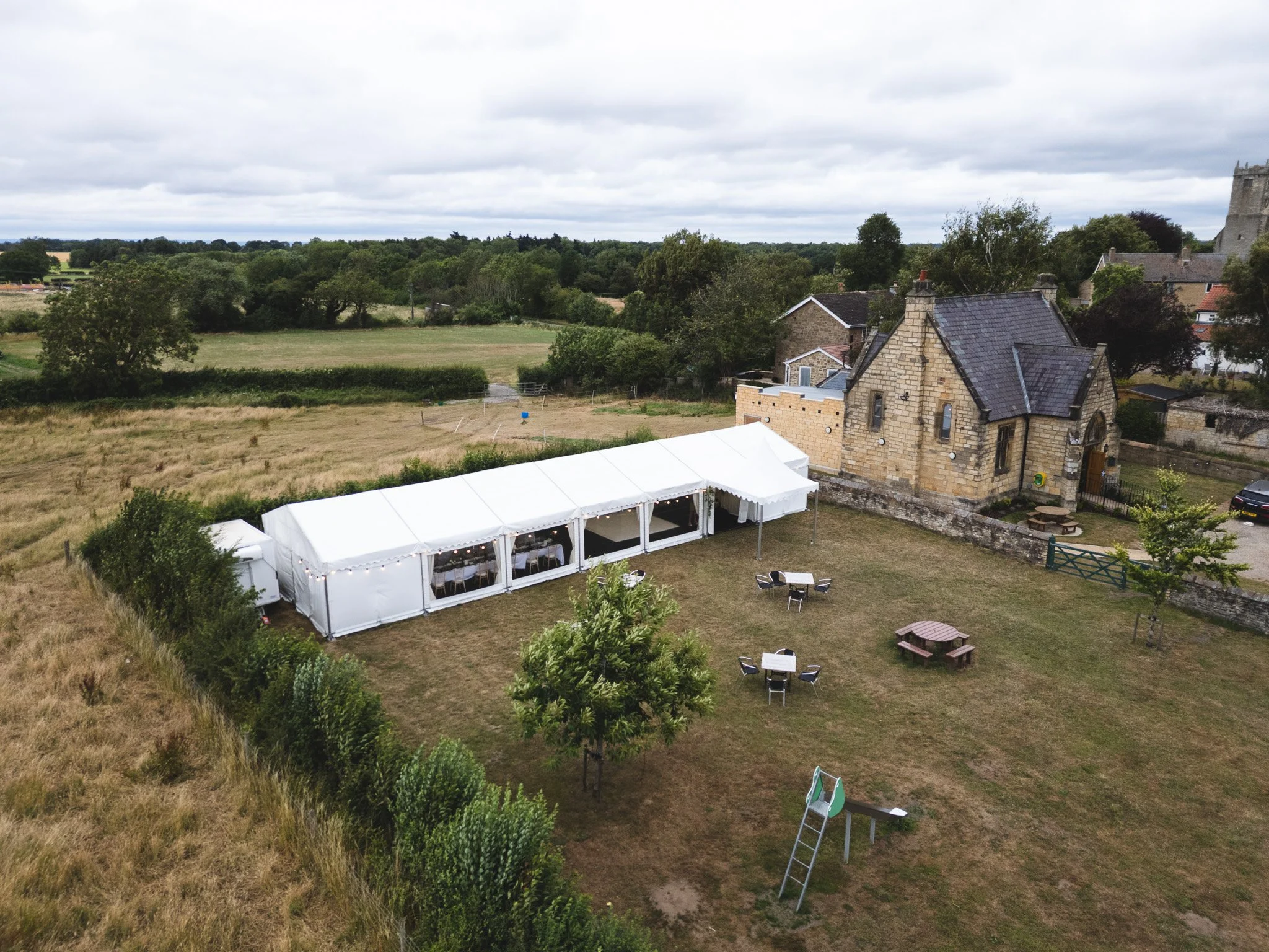 A large outdoor tent set up on a grassy area near a historic stone building with a slate roof. The scene includes scattered picnic tables, chairs, and a ladder, with trees and open fields in the background under a cloudy sky.