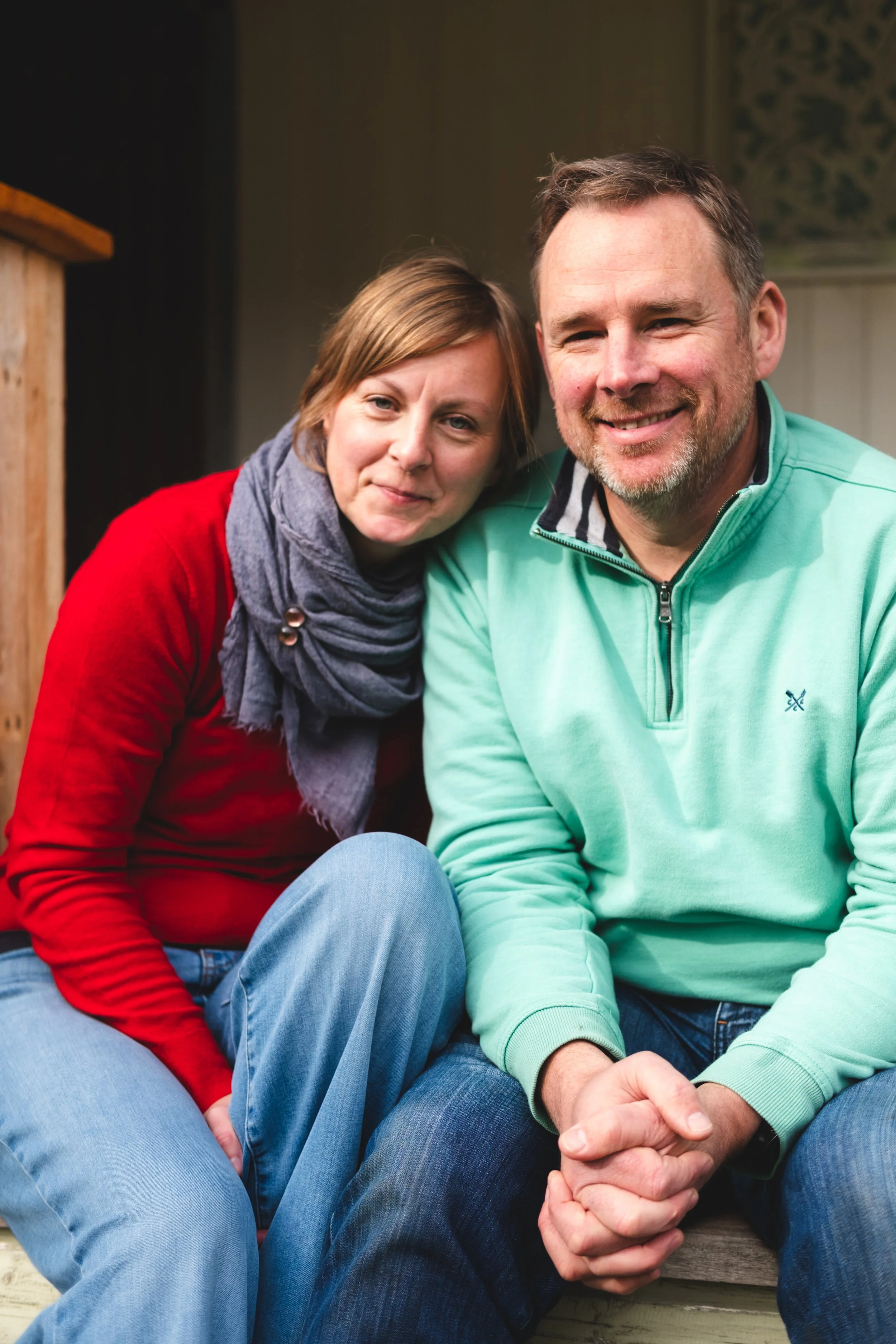 A smiling couple sitting close together, with the woman leaning on the man's shoulder, outdoors or in a cozy indoor setting.