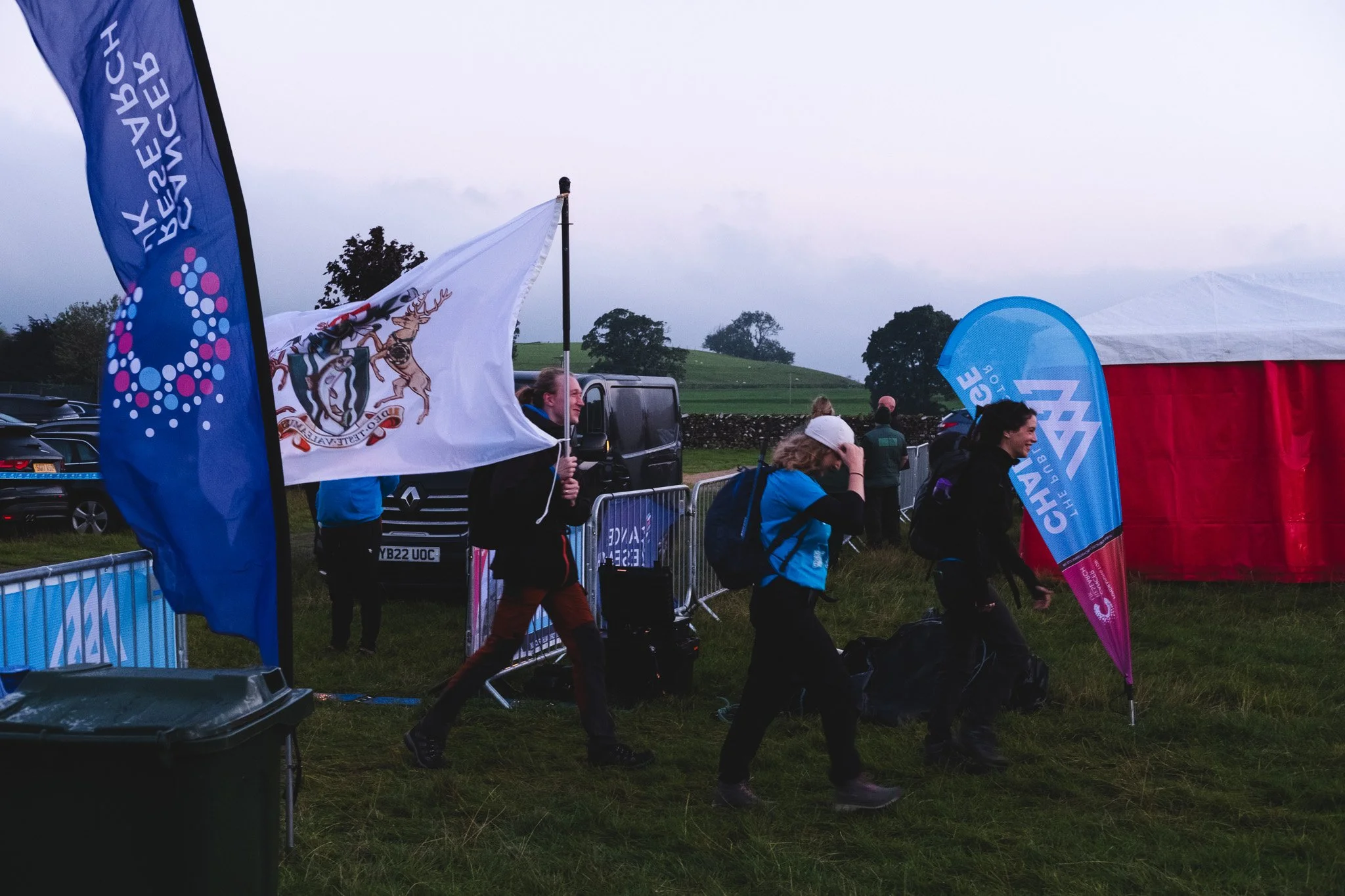 People walking past flags and barriers at an outdoor event in a grassy field, with a tent, parked cars, and trees in the background on a cloudy day.