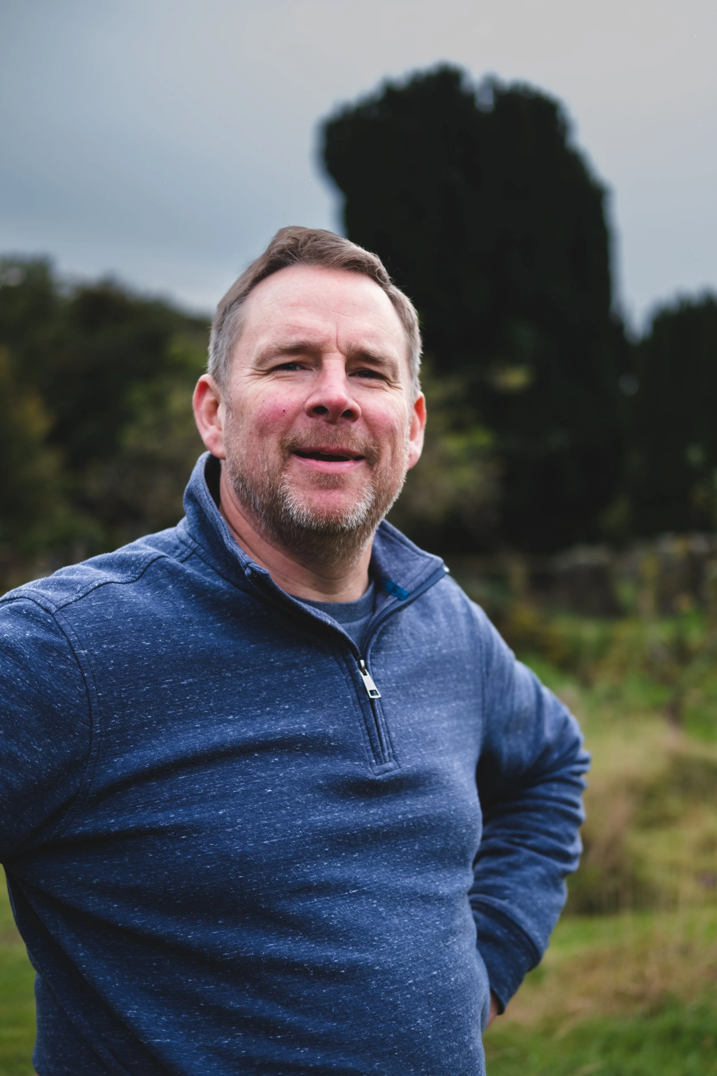 A middle-aged man with short brown hair, a beard, and light skin, standing outdoors in a rural area with green trees and overcast sky in the background. He is wearing a blue zip-up sweater and smiling at the camera.