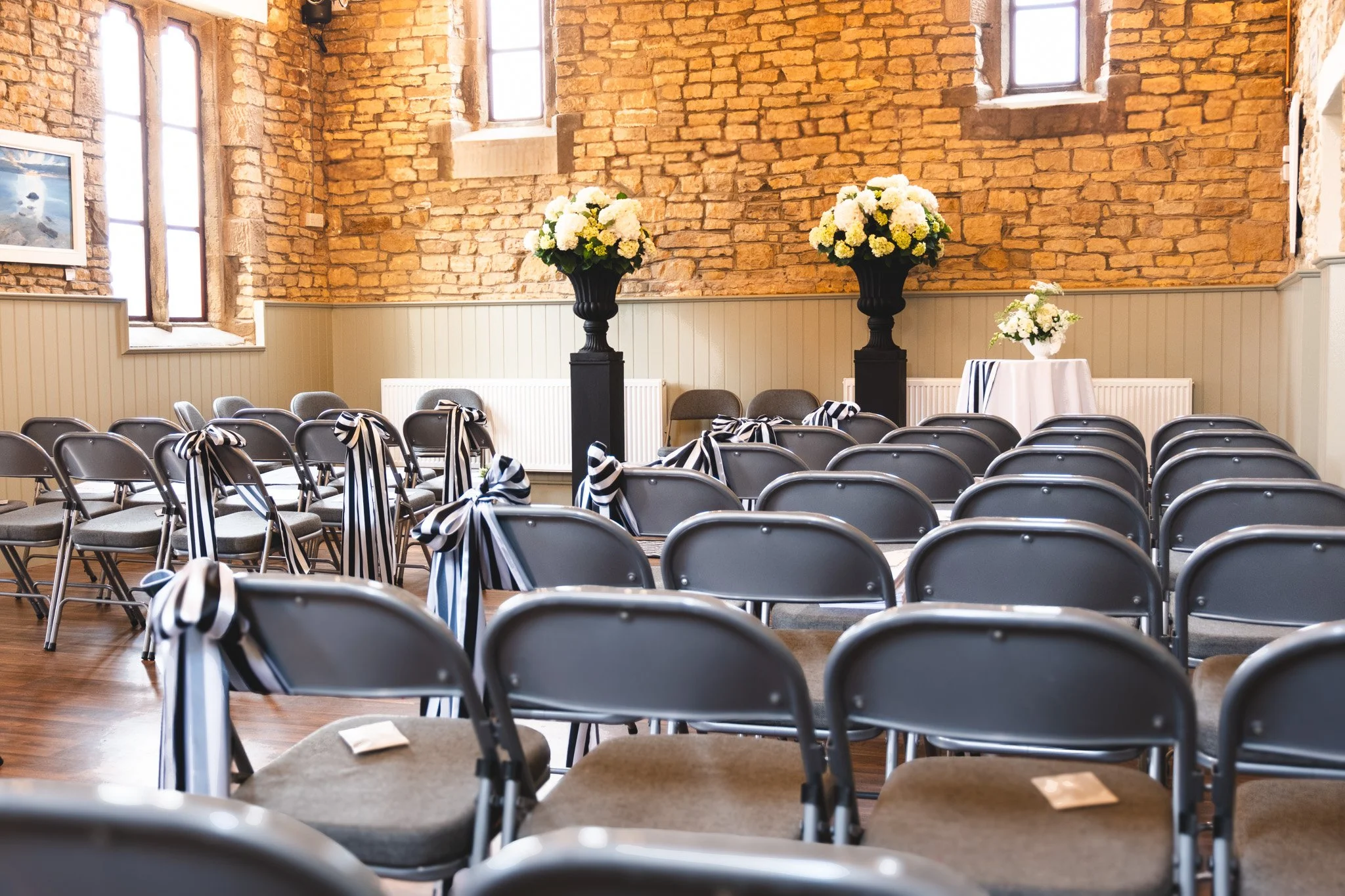 Indoor wedding or event ceremony setup with rows of gray folding chairs decorated with black and white striped sashes, facing a small table and large flower arrangements with white flowers in black vases against a rustic brick wall background.