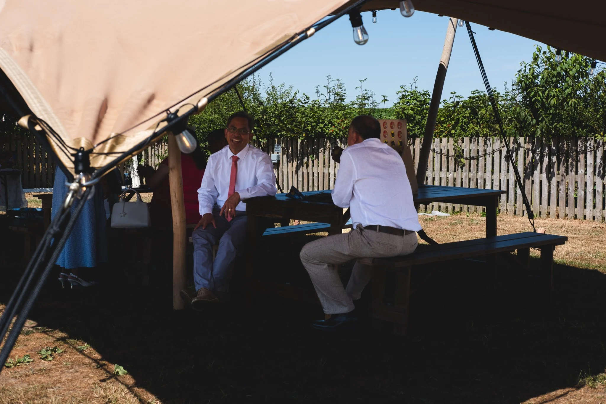 People sitting under a large canopy outdoors, engaging in conversation, with a wooden fence and greenery in the background.