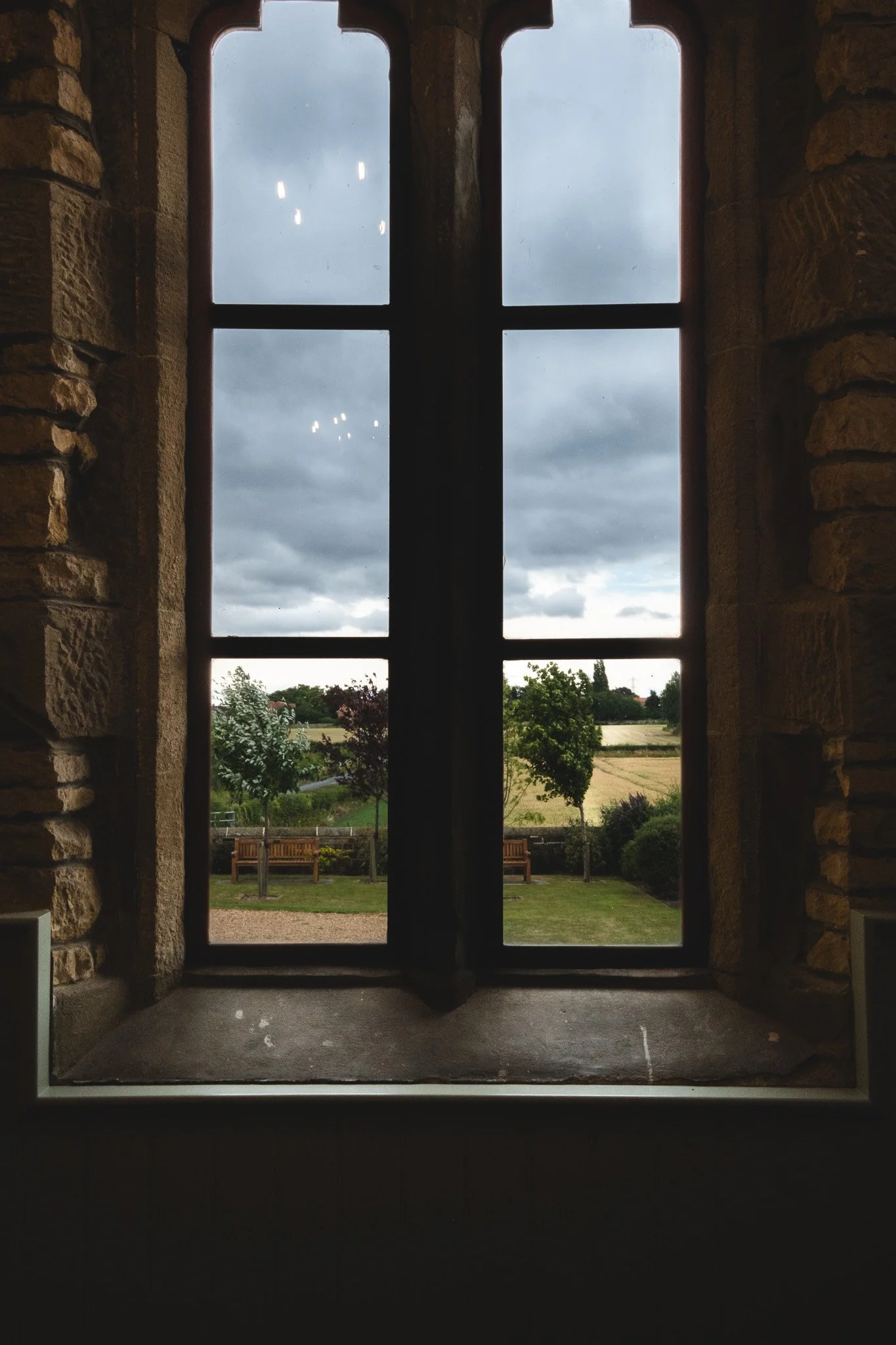 View of a cloudy sky and outdoor landscape through a tall, arched stone window with metal framing.