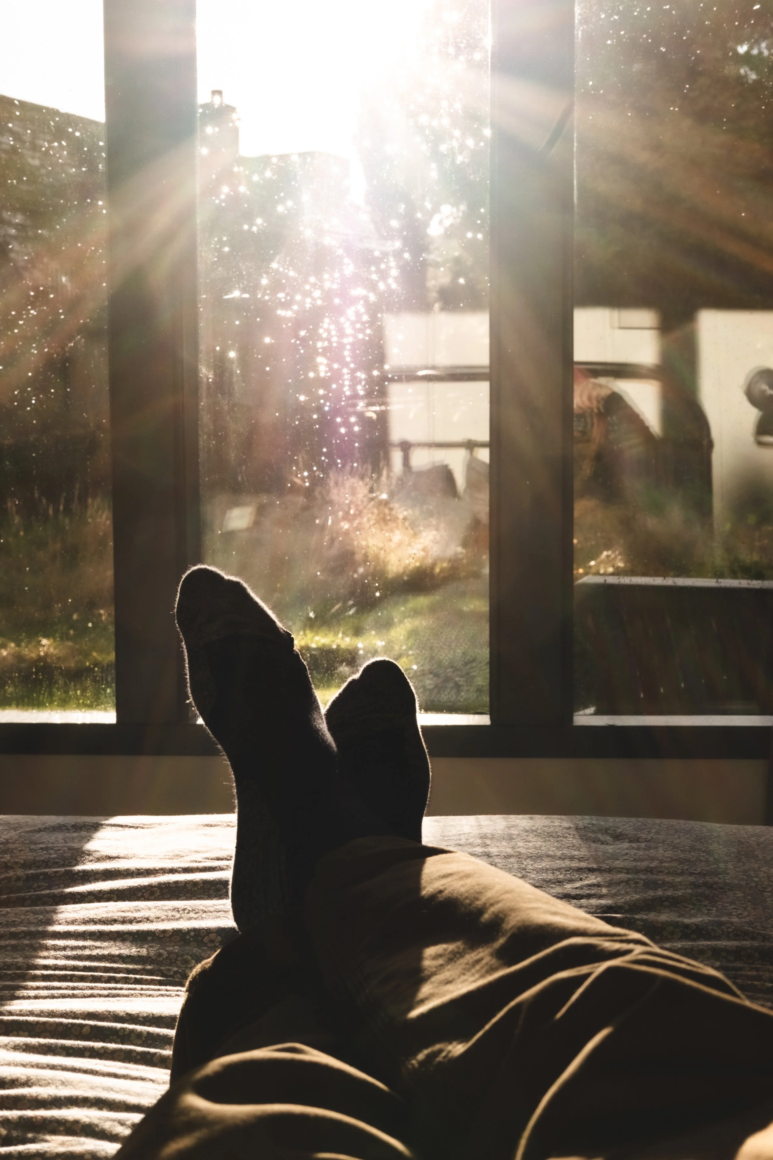 Person lounging on a bed with feet up, looking out a sunlit window in the late afternoon, with sunlight creating a sparkling effect on the glass.