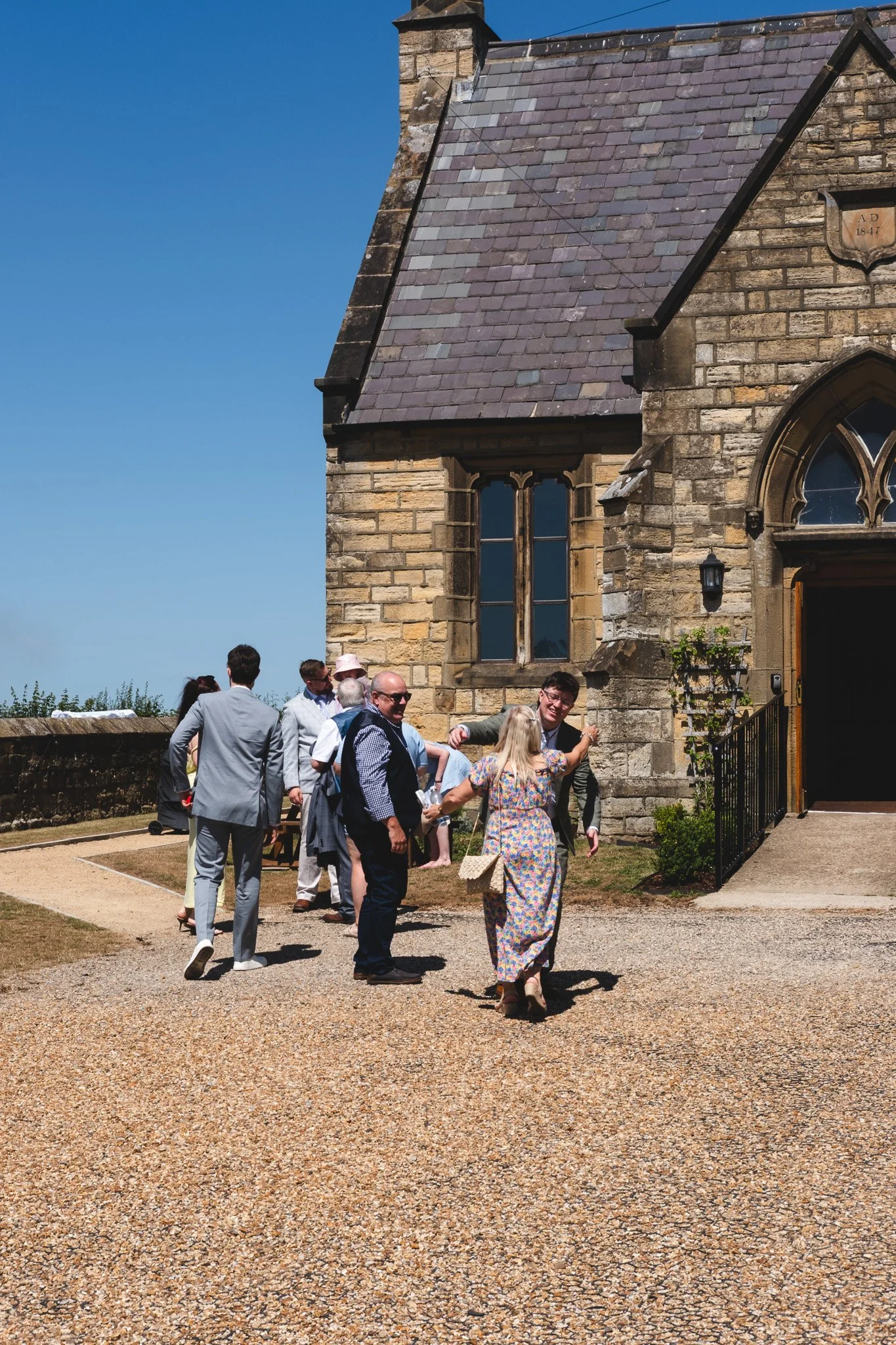 A group of people dressed in formal and casual attire standing outside a historic stone church, some talking and interacting on a sunny day with clear blue sky.