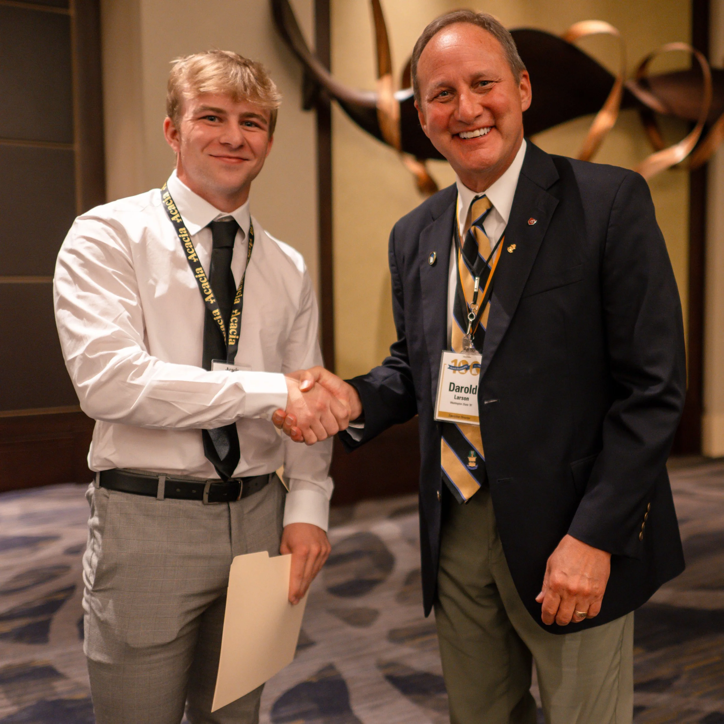 Two men in suits shake hands on stage during an event, with a banner in the background reading 'William A. Utic, Acacia Fraternity Foundation, Scholarship Banquet' and a man holding a clipboard.