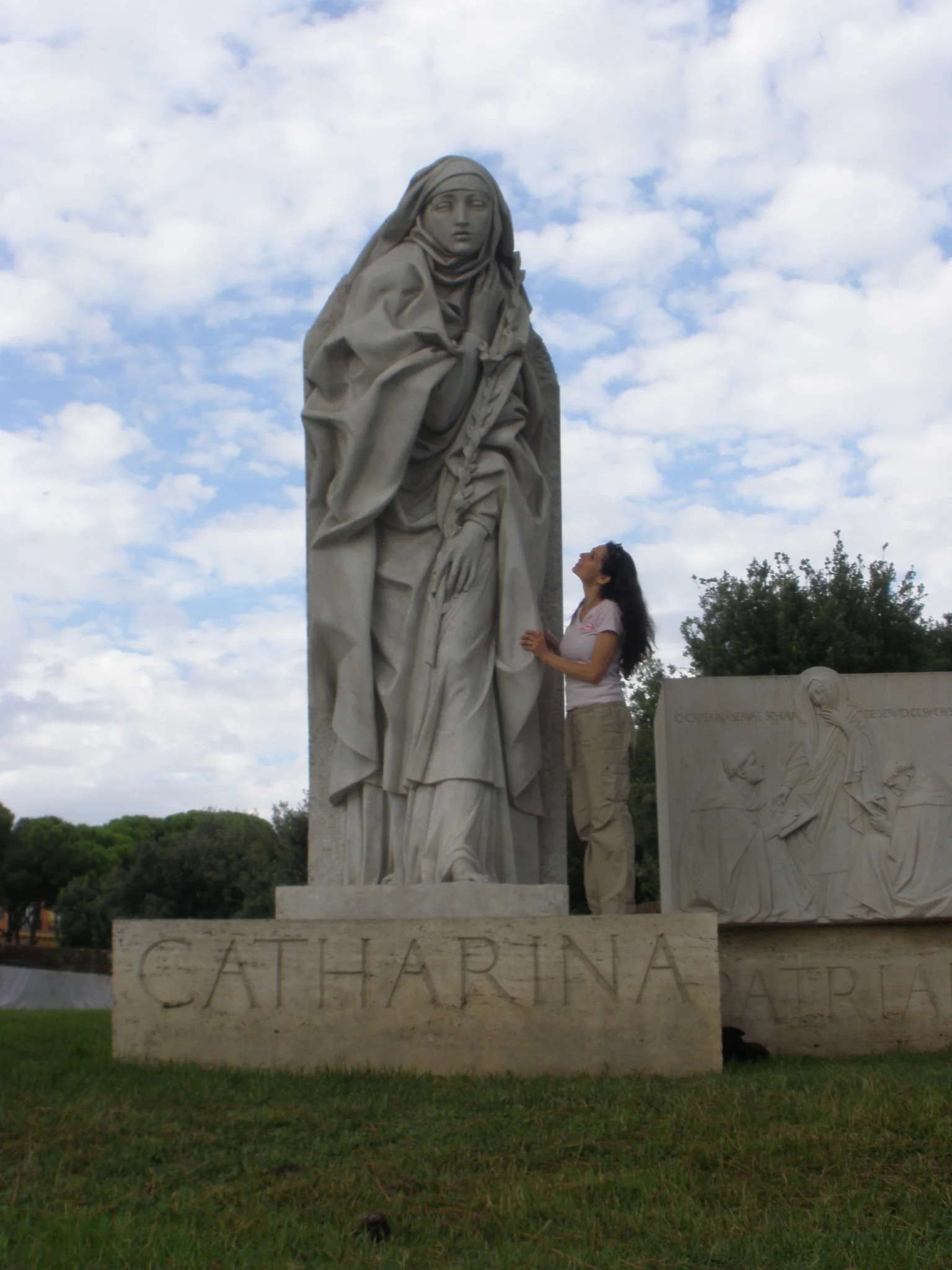 A woman standing next to a large stone statue of a woman in a cloak with the word 'CATHARINA' carved on the base, in an outdoor park with trees and a partly cloudy sky.