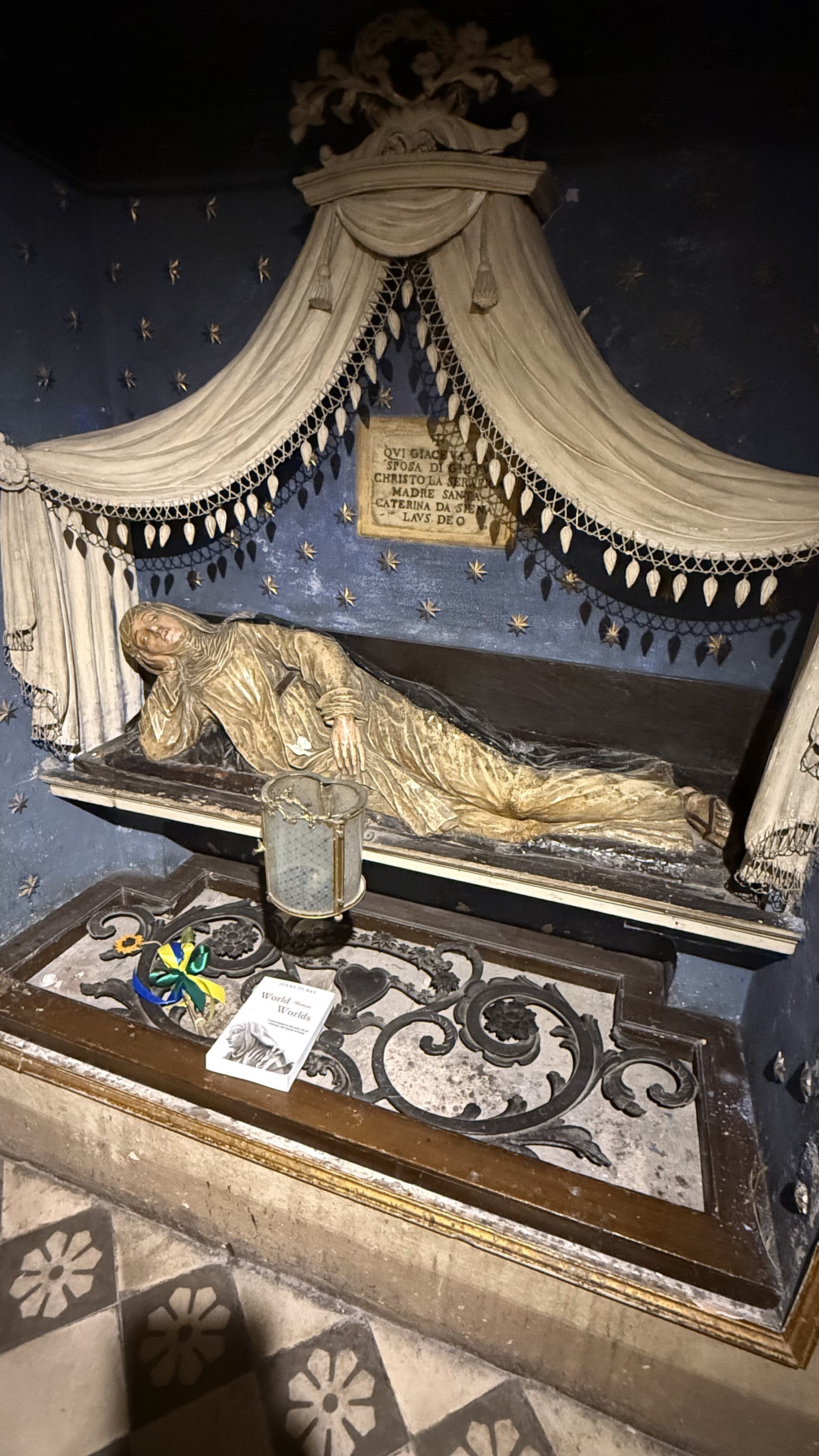 Statue depicting a reclining figure of St. Catherine of Siena, with decorative drapes and stars in the background, likely part of a religious display.