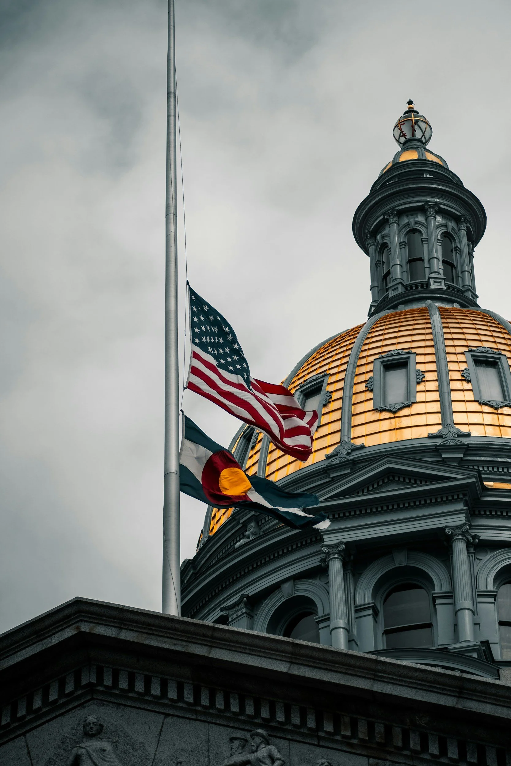 The Colorado State Capitol building with American and Colorado flags flying in front, and a cloudy sky overhead.
