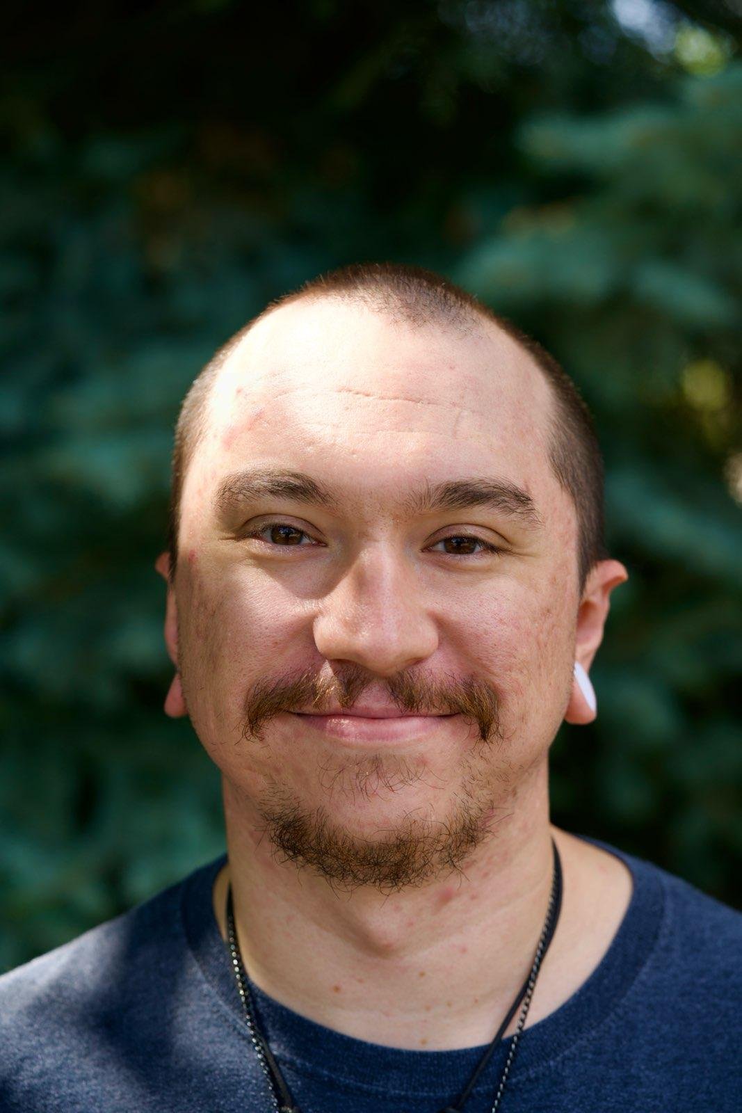 Close-up of a young man with a mustache and beard, smiling, with a blurred green foliage background.