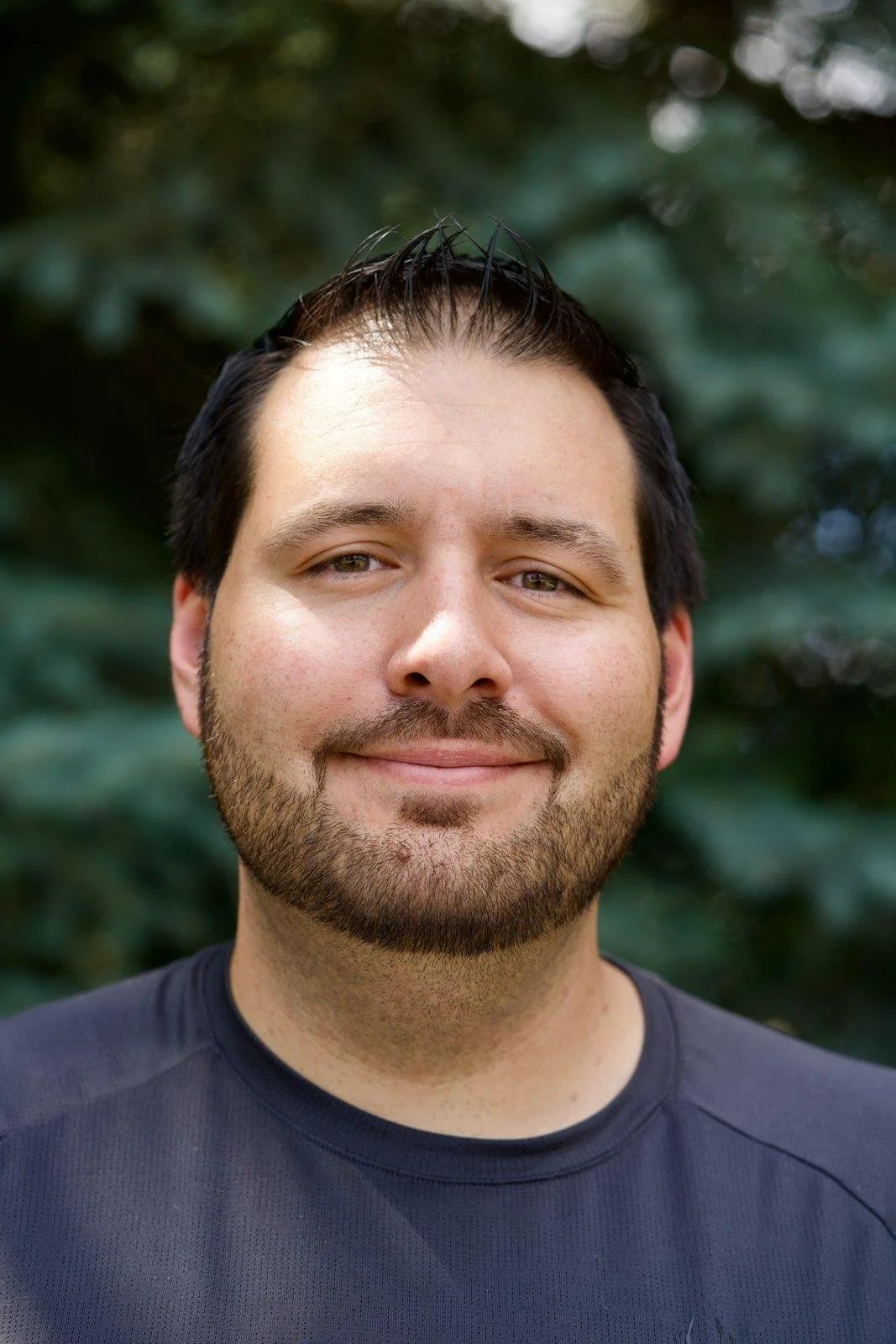 A close-up portrait of a smiling man with short dark hair, a beard, and light-colored eyes, wearing a dark shirt, outdoors with blurred green foliage in the background.