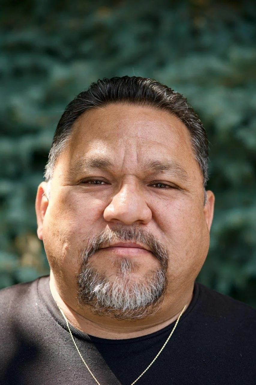 Close-up portrait of a middle-aged man with a mustache and goatee, short black and gray hair, wearing a black shirt and a gold chain, standing outdoors with green foliage in the background.