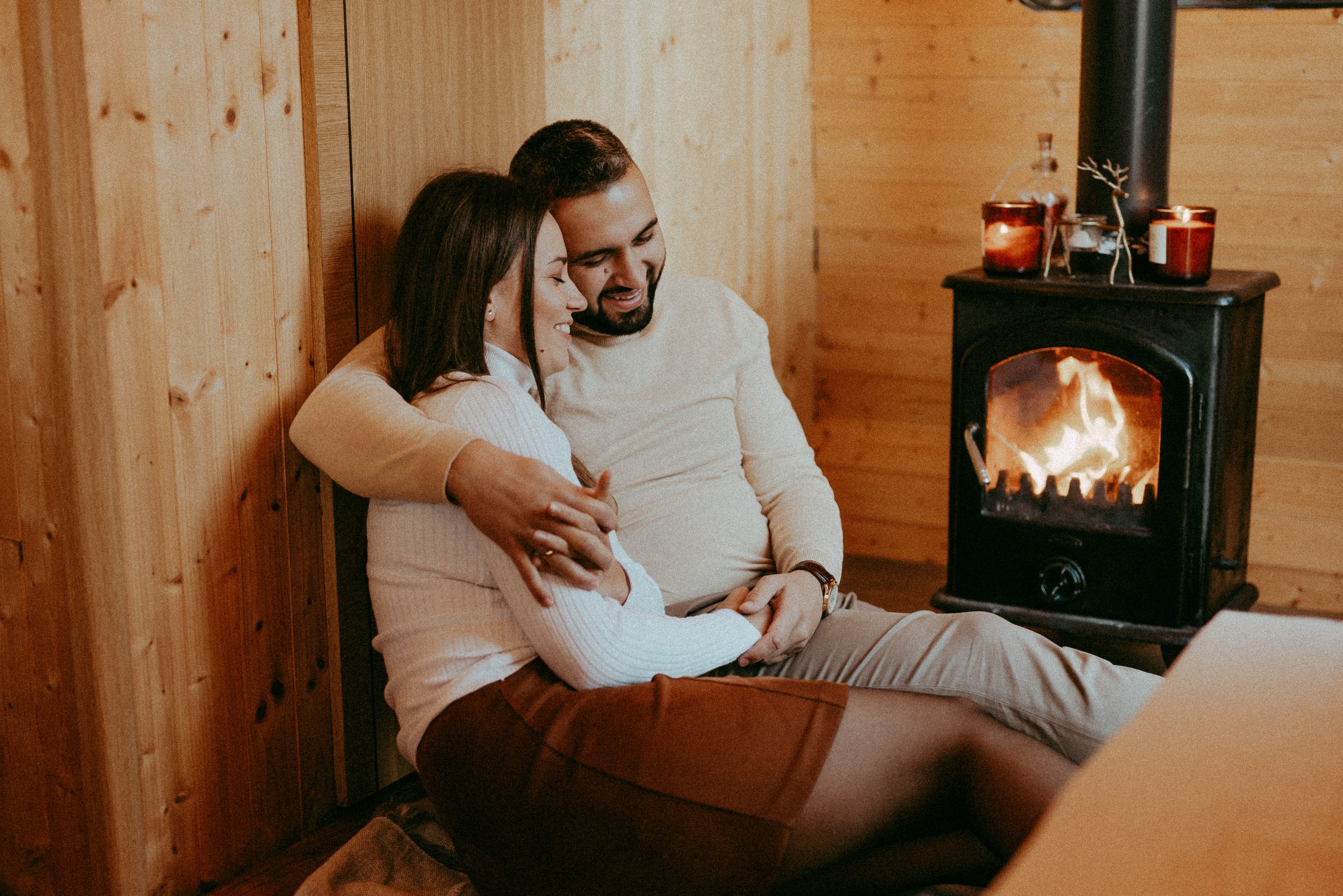 A couple sitting close together near a fire in a cozy, wood-paneled room, smiling and embracing.