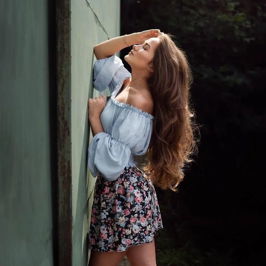 A young woman with long wavy hair leaning against a mossy wall outdoors, with her eyes closed and one hand resting on her forehead.