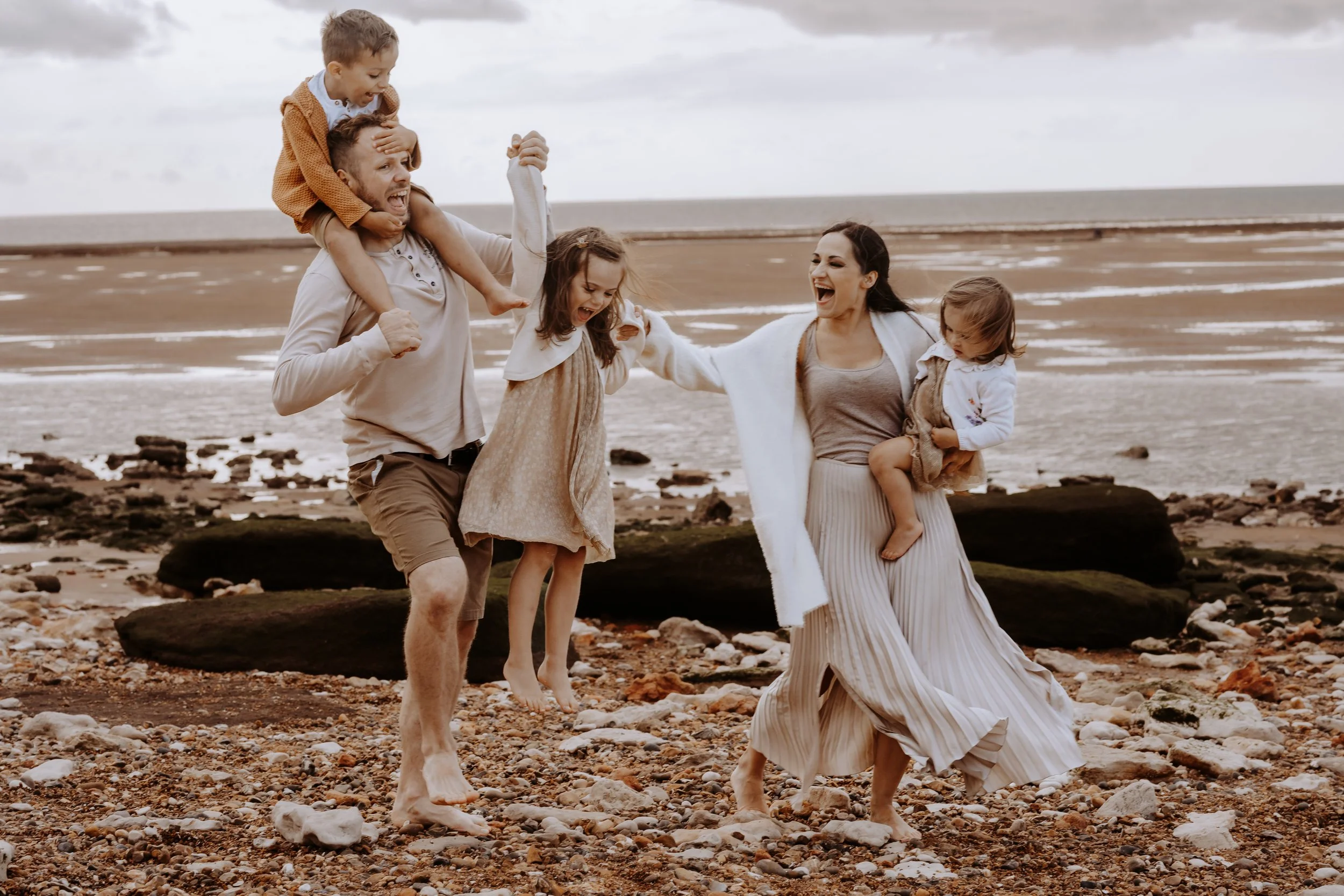 A group of five people, two adults and three children, enjoying a day at the beach. One man carries a young boy on his shoulders, while another woman holds a young girl. The group is smiling and laughing on a rocky, sandy beach with the ocean in the 