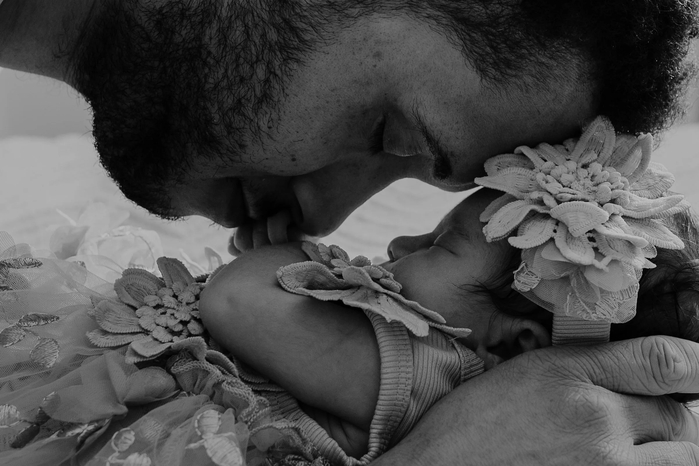 A man gently kisses a sleeping baby on the forehead, both wearing floral headbands, in a black and white photo.