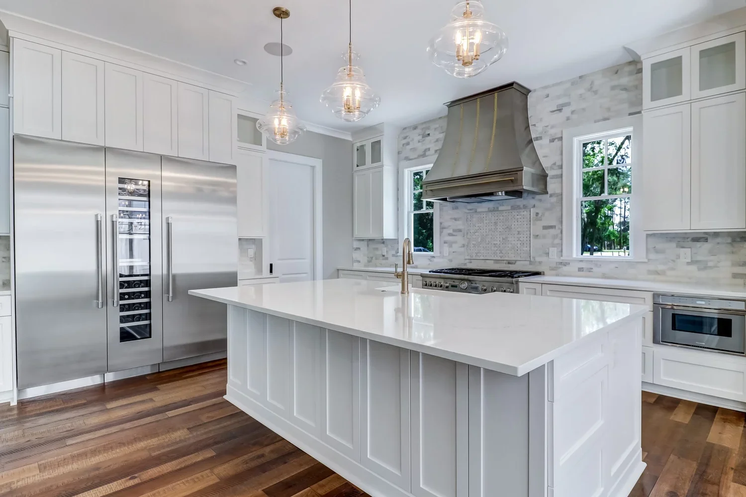 Modern white kitchen with stainless steel refrigerator, island with white countertop, gold faucet, and a range with a vent hood, wooden flooring, and two windows showing green trees outside.
