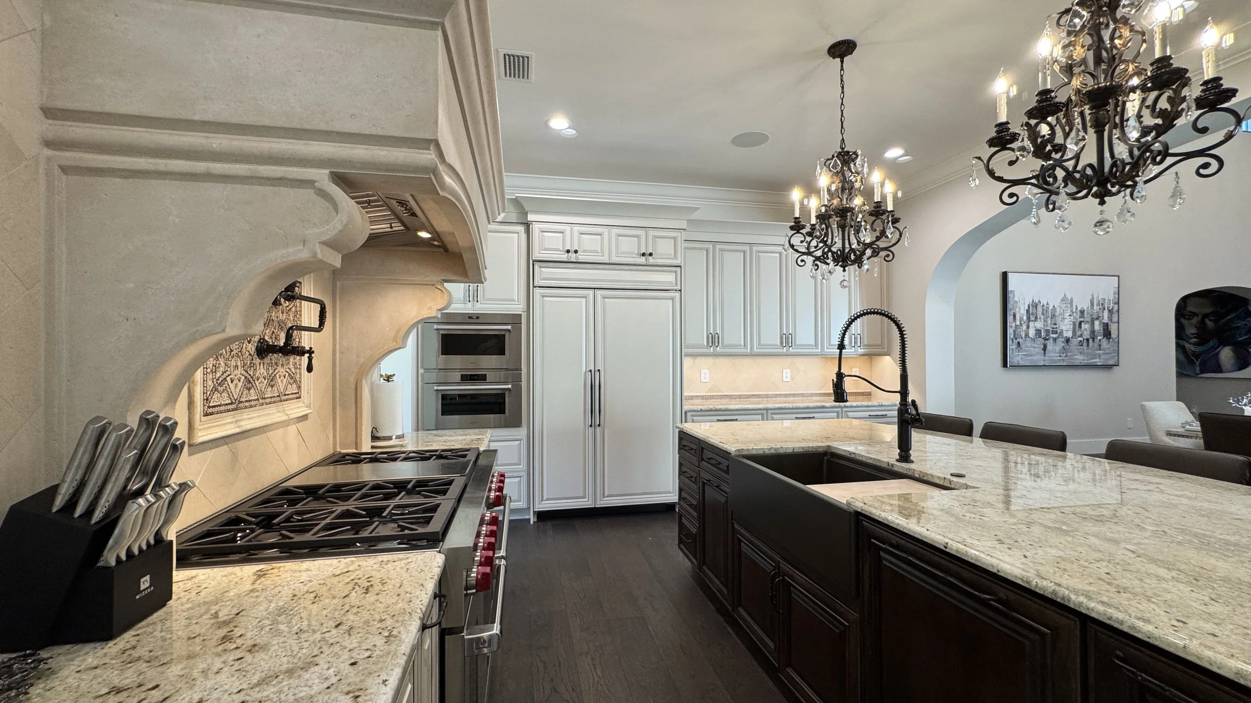 Modern kitchen with white cabinetry, granite countertops, dark island, stainless steel appliances, and ornate chandelier lighting.