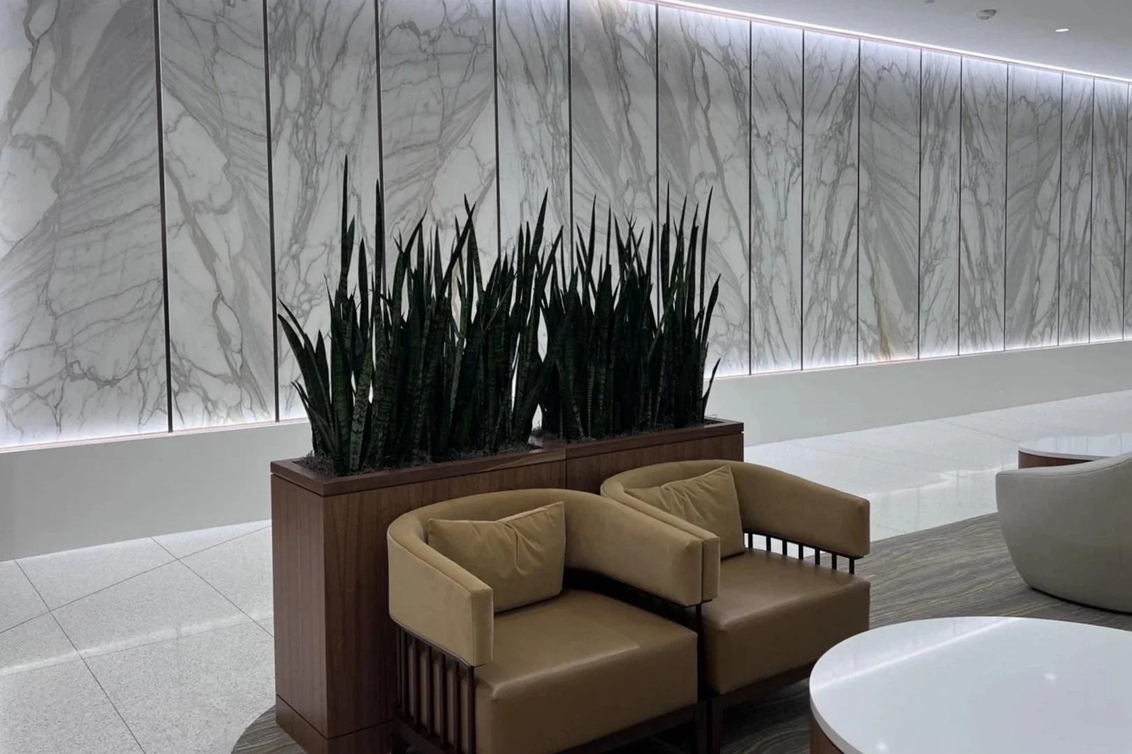 Modern lobby area with beige couches, a white round table, and a large marble wall with backlighting. There are tall indoor plants in a wooden planter.