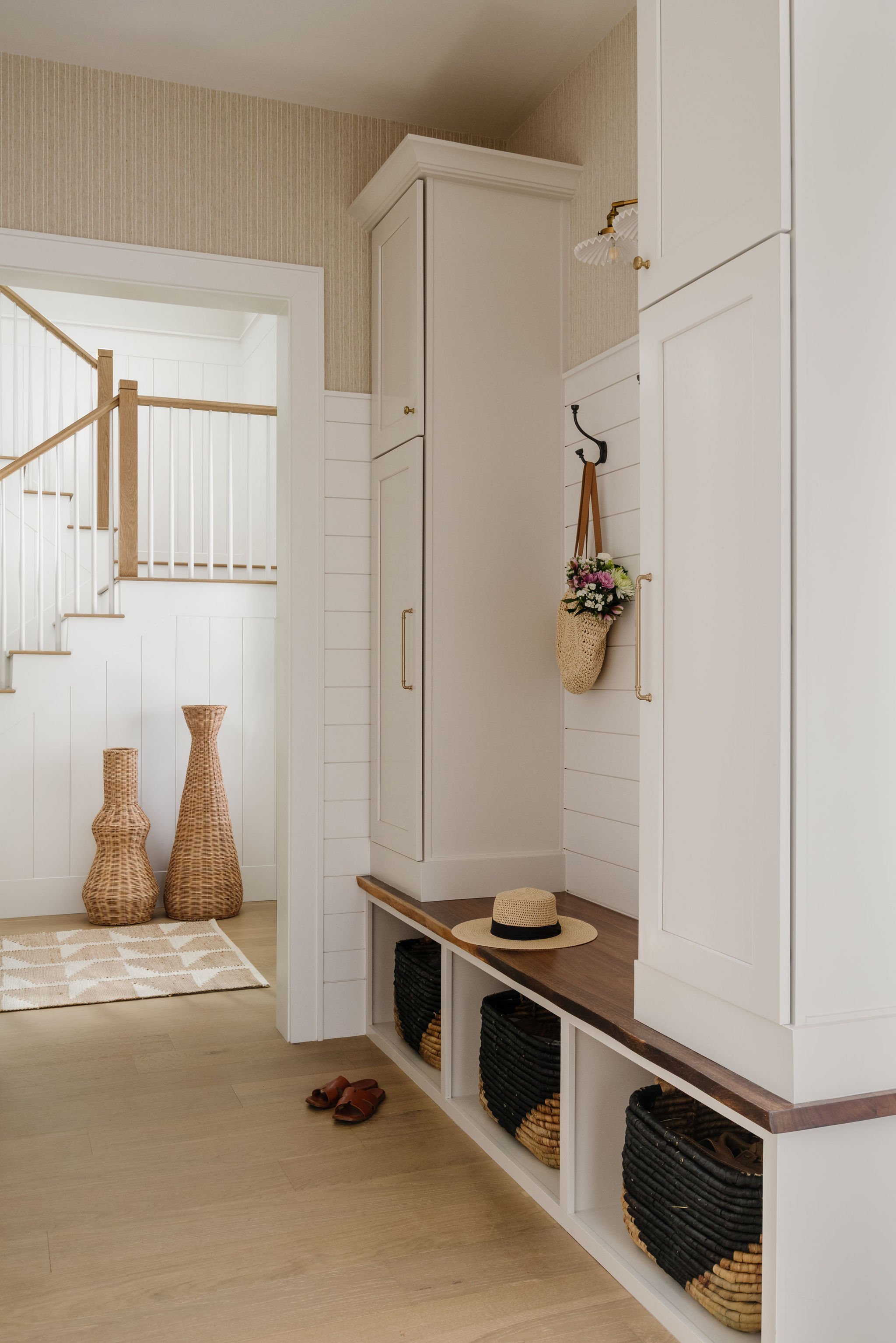 Entryway with white built-in storage, baskets, a hat, and slippers, and stairs with wooden handrails in the background.