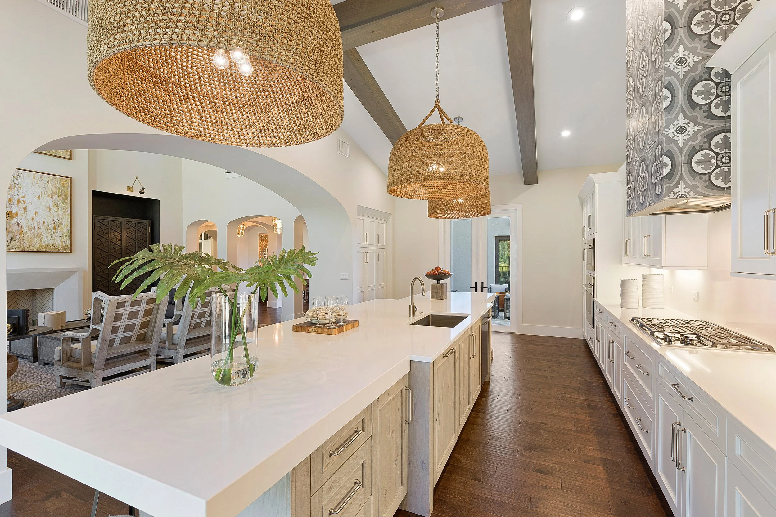 A modern kitchen with white cabinets, a large central island, and three woven pendant lights hanging from the ceiling. There is a stove on the right, a faucet and sink on the island, and a decorative wallpapered section near the stove. A dining area with chairs and a large plant are visible in the background.