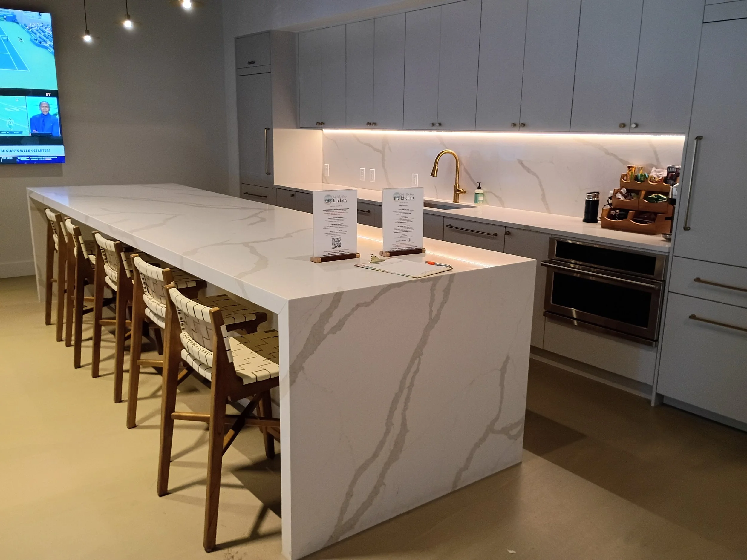 Modern kitchen with white marble island, grey cabinets, a gold faucet, and a wall-mounted TV on the left showing sports news.