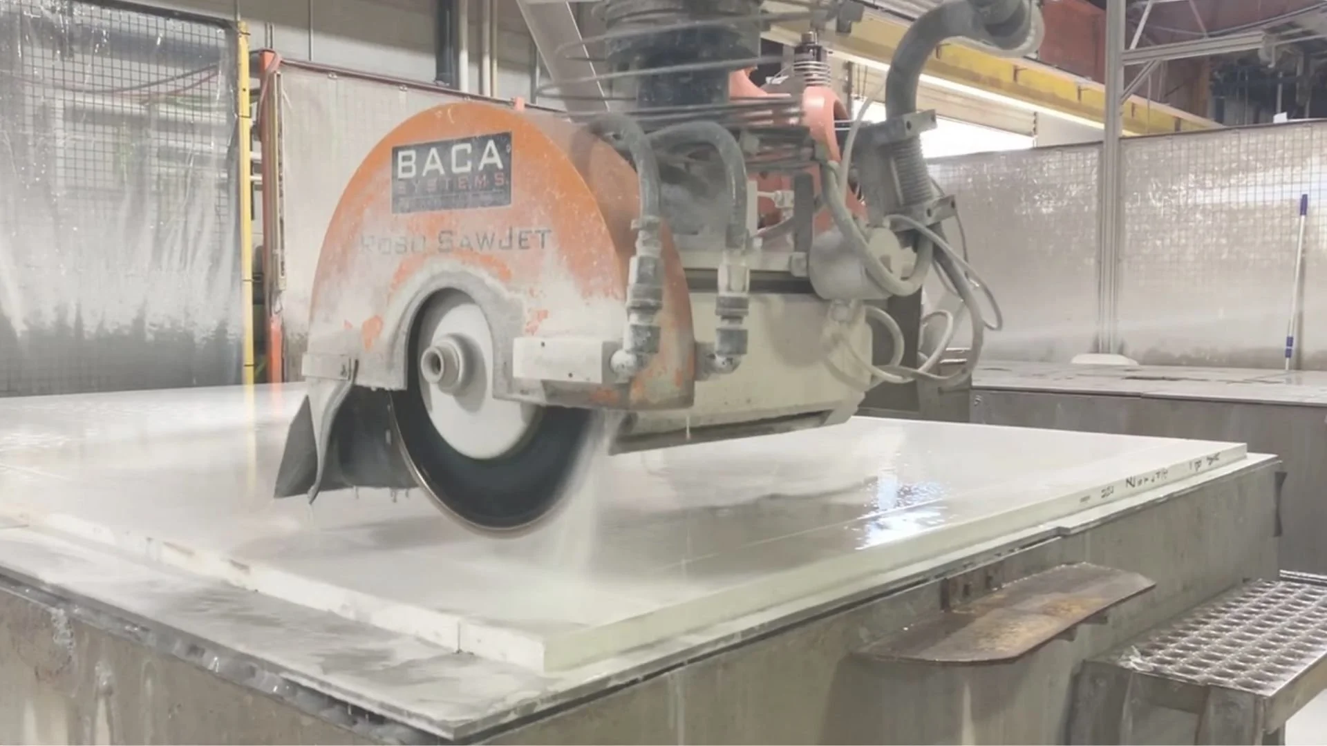 A large industrial circular saw cutting through a large slab of material, with dust and debris in the air, in a factory or workshop environment.
