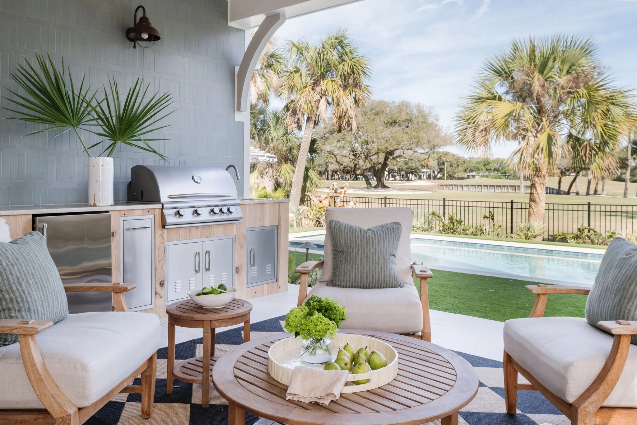 Outdoor patio area with white cushioned chairs, a round wooden coffee table with a bowl of green apples, and a small side table with green flowers. In the background, there's a built-in outdoor kitchen with a grill and a large plant in a white vase. 
