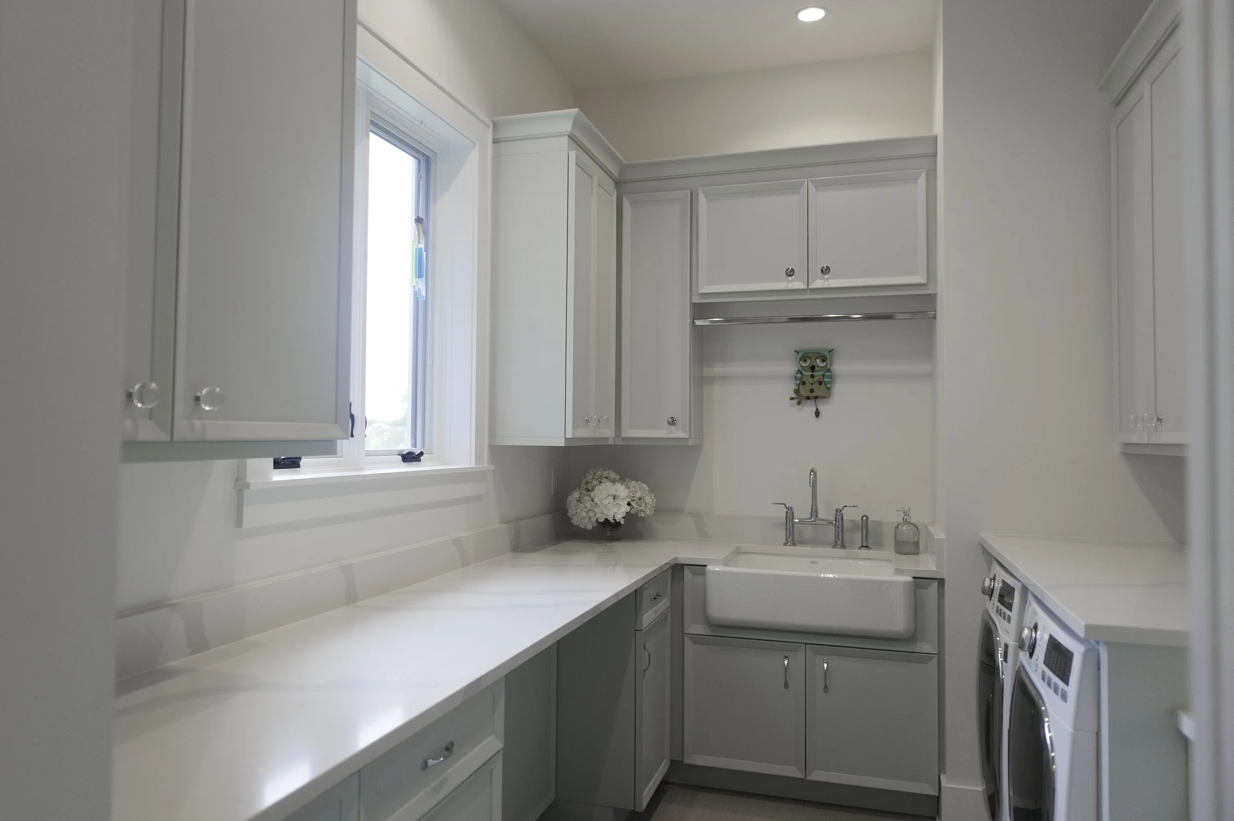 A bright, modern kitchen with white cabinets, a white countertop, and a window letting in natural light. There are flowers on the counter and a small owl-shaped decorative item on the wall.