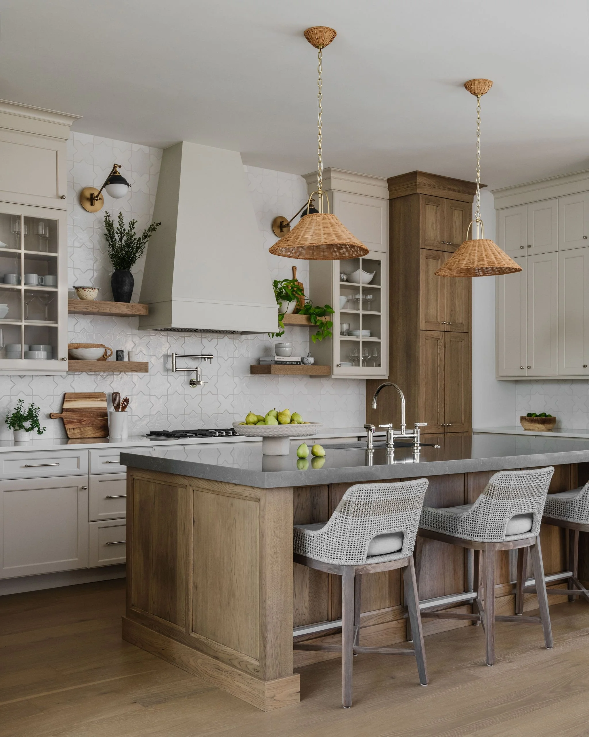 Modern kitchen with white cabinets, wooden accent elements, a kitchen island with a gray countertop, two wicker pendant lights, and bar stools with woven backs