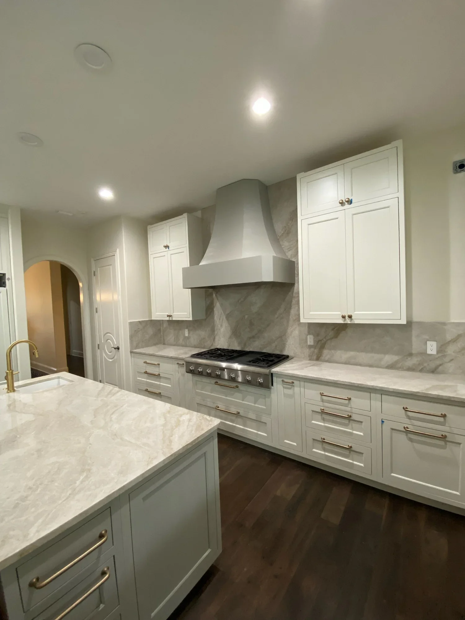A modern kitchen with white cabinets, a marble countertop, a stove, and a marble backsplash. There is a large island with a marble surface in the foreground, and a stainless steel stove with a vent hood in the background. The kitchen has dark hardwood floors.