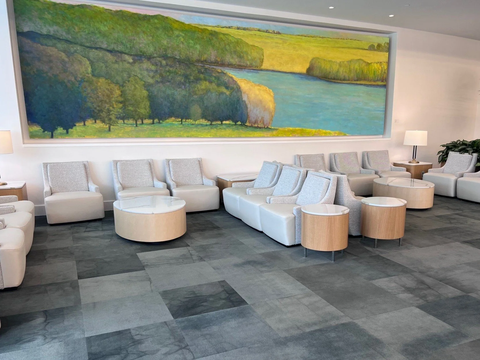 A modern waiting area or lounge with beige armchairs and round wooden coffee tables. There is a large landscape painting of a river, trees, and grassy fields on the wall behind the seating. Floor lamps and potted plants are also visible.