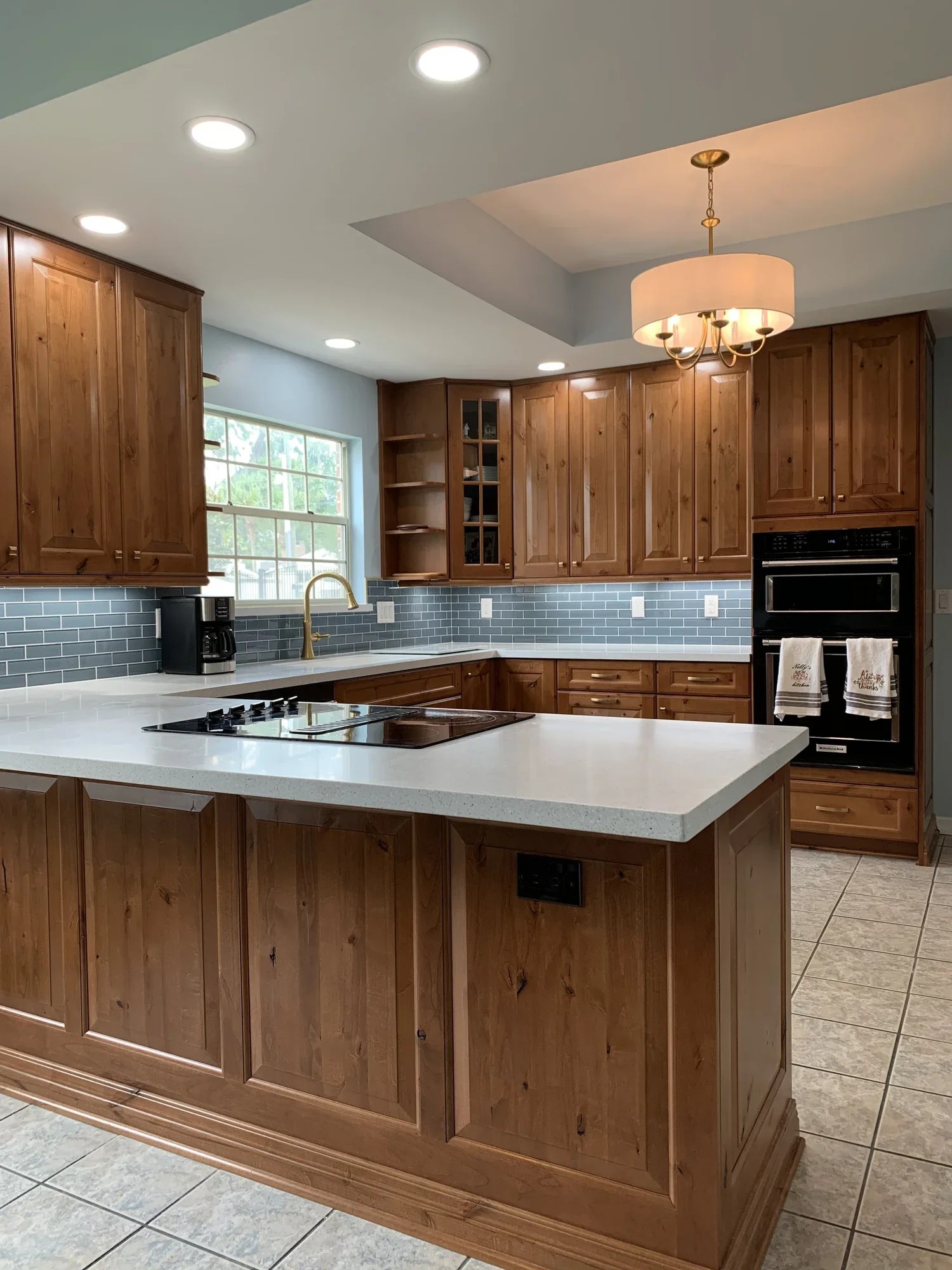 A kitchen with wooden cabinets, a white countertop, a window, a black coffee maker, a gold faucet, recessed lighting in the ceiling, and a hanging light fixture over the counter. There are also paper towels hanging on the oven handle.