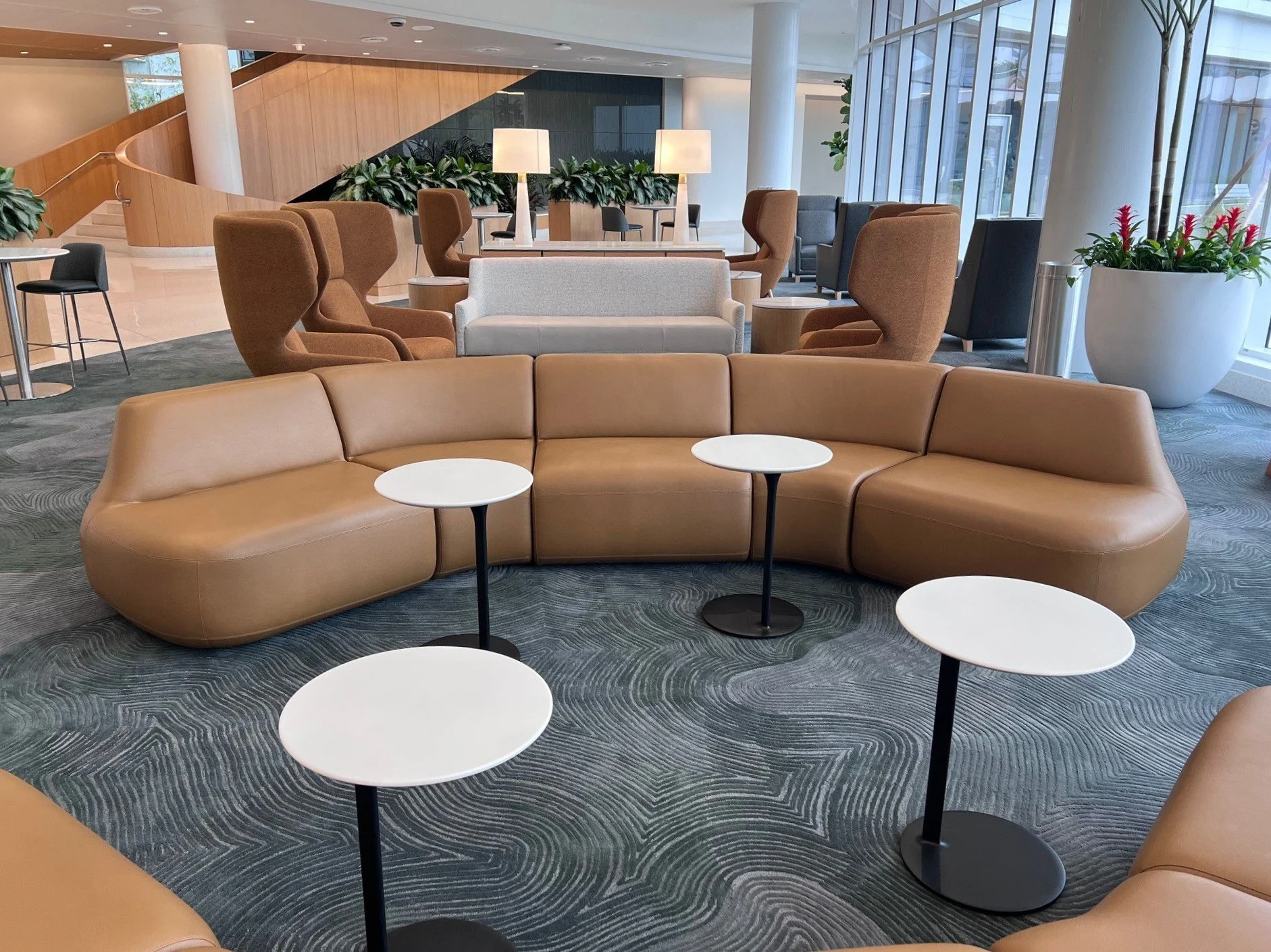 Lobby area with a curved tan sofa, small round black and white tables, brown and gray armchairs, tall white lamps, and large windows with greenery outside.