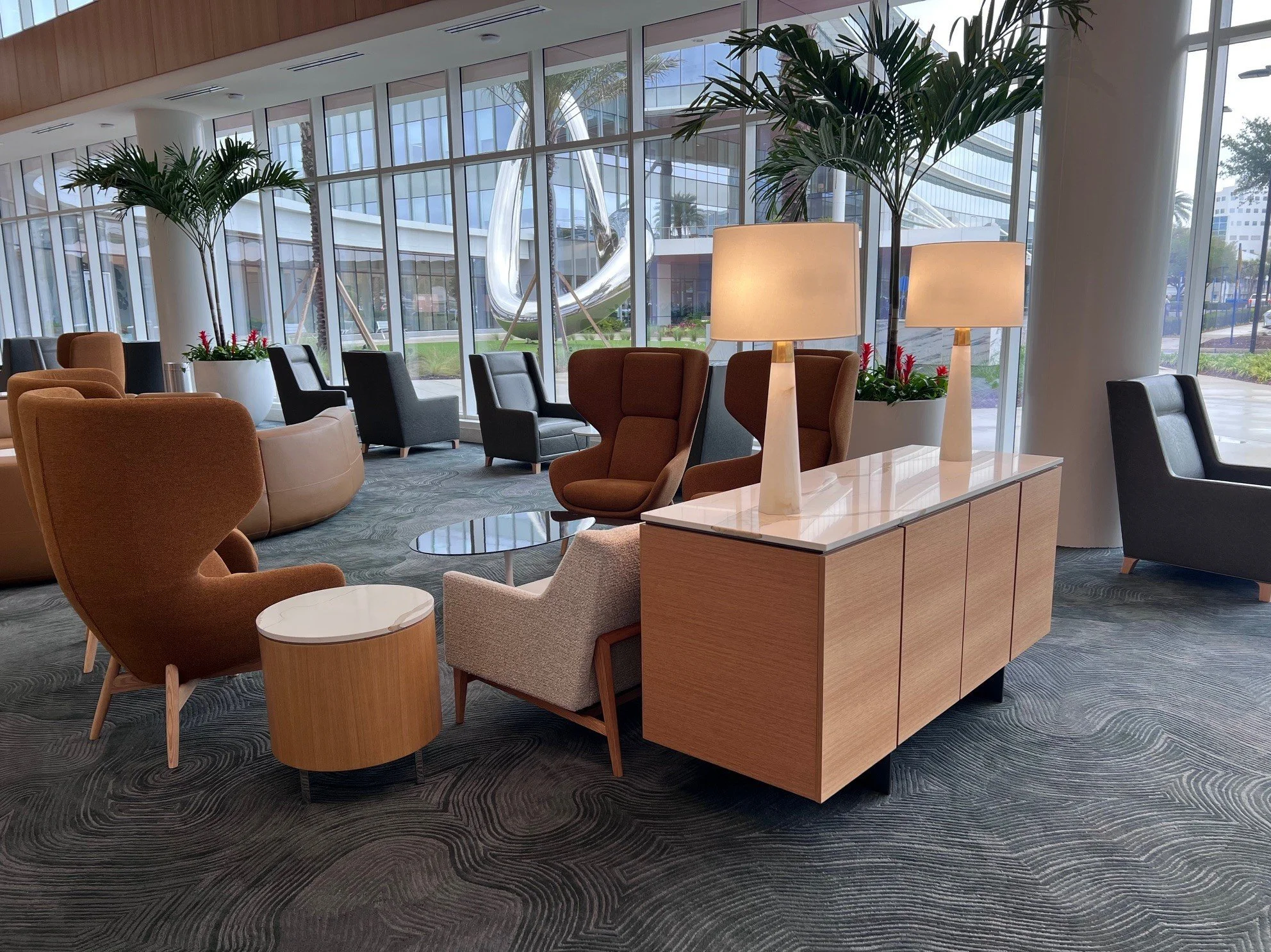 Interior of a modern lounge area with multiple colorful chairs, wooden furniture, large decorative lamps, and large windows with views of trees and a building outside.