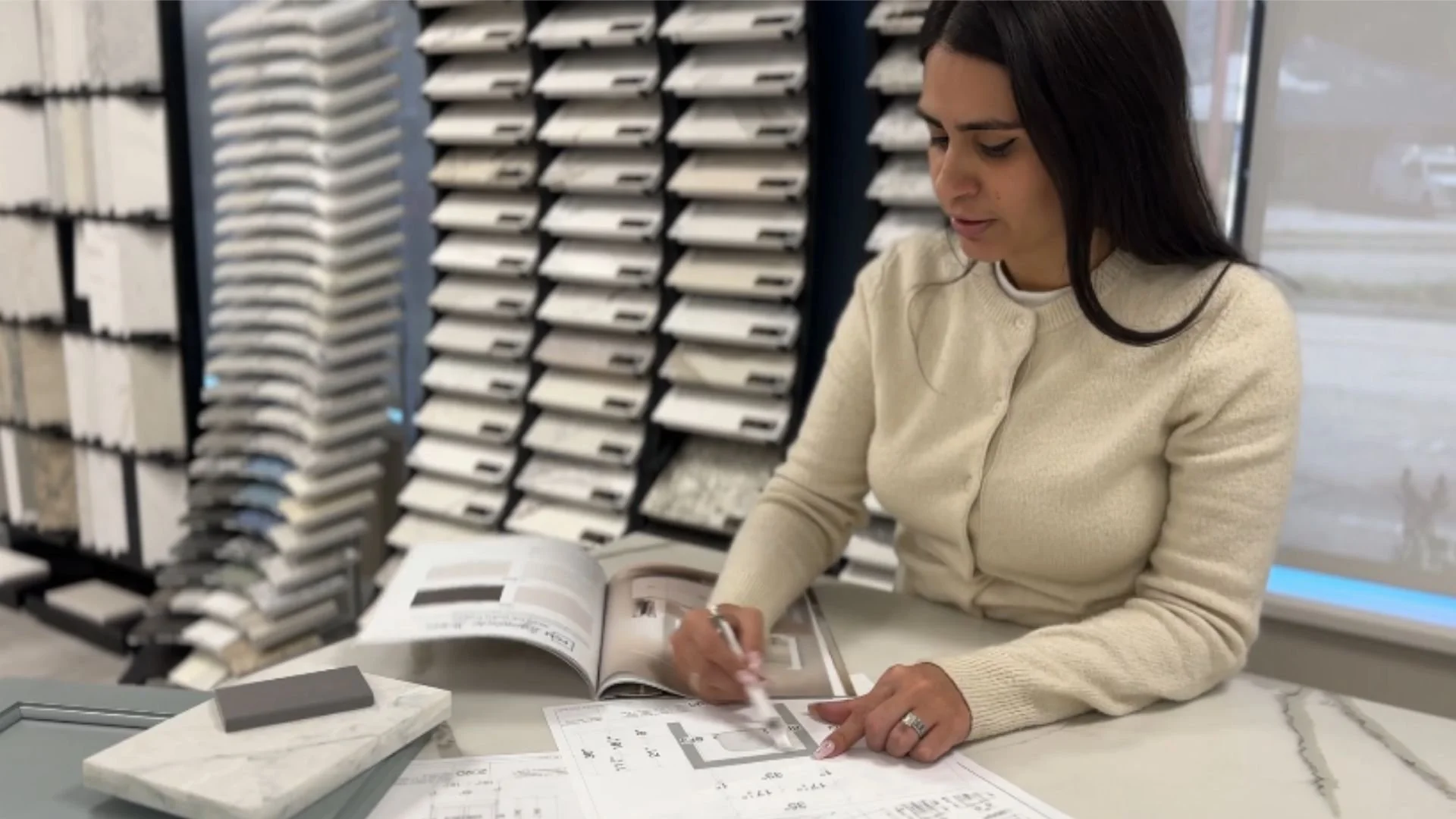 Woman sitting at a table, examining design samples and reading a catalog in a showroom with color swatches and material samples.