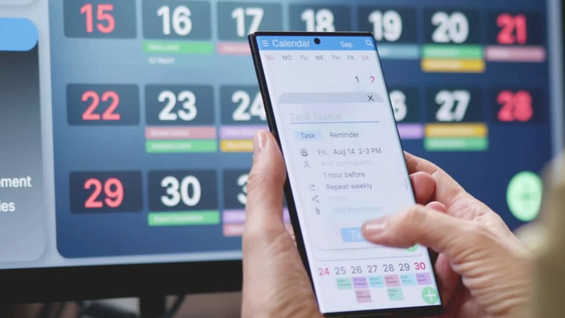 A person holding a smartphone displaying a calendar app with scheduled tasks and reminders, in front of a large digital calendar on a screen showing dates in various colors.
