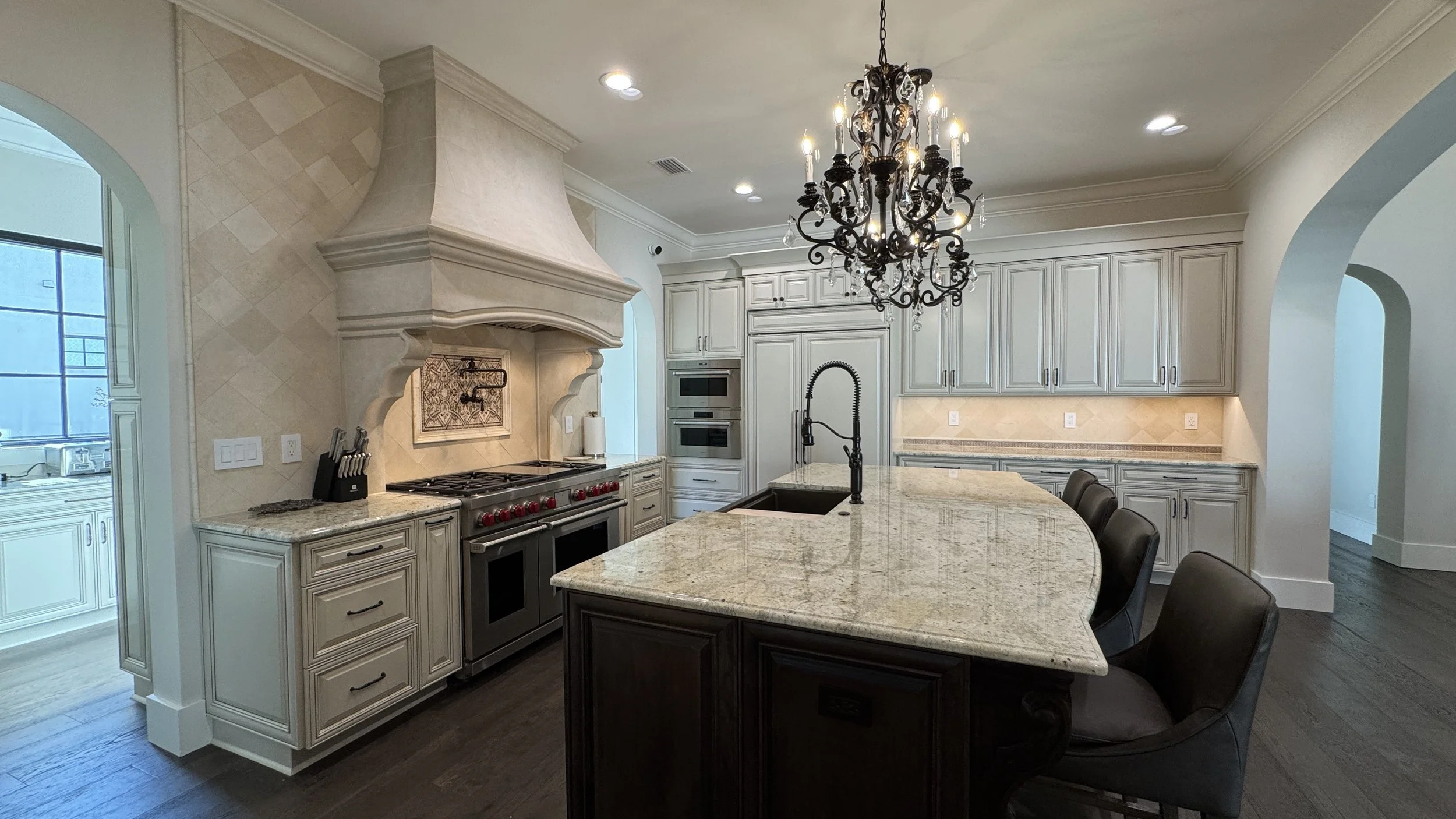 A spacious kitchen with cream-colored cabinets, a large island with a granite countertop, a built-in oven, and a chandelier hanging above the island.