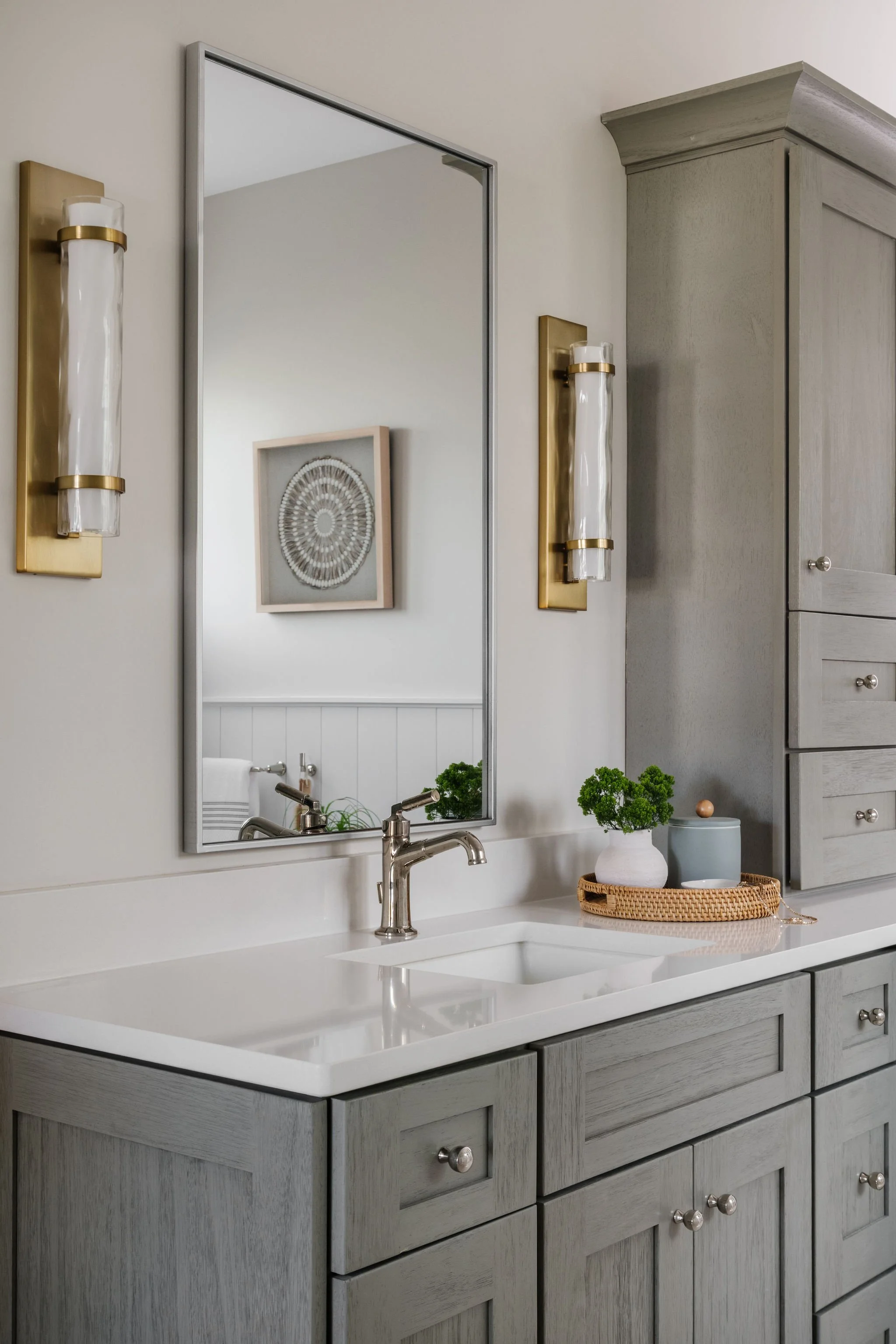 A bathroom vanity with a white countertop, gray wood cabinetry, a rectangular mirror, and decorative wall sconces with gold bases. There are two small potted plants and a tray with accessories on the countertop.