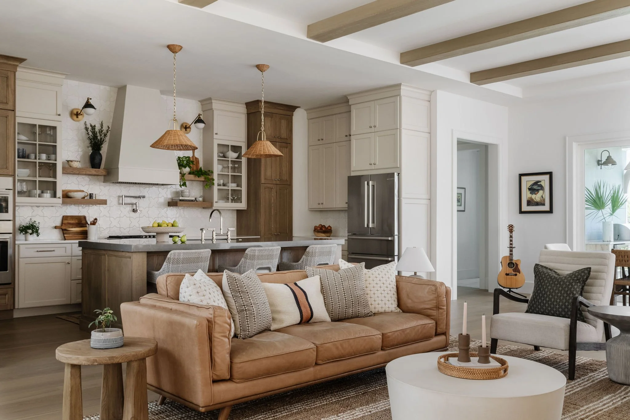 Open-concept living room and kitchen with neutral tones, wooden accents, and modern decor, including a leather sofa, patterned pillows, a round coffee table, and kitchen cabinets with open shelves and pendant lighting.